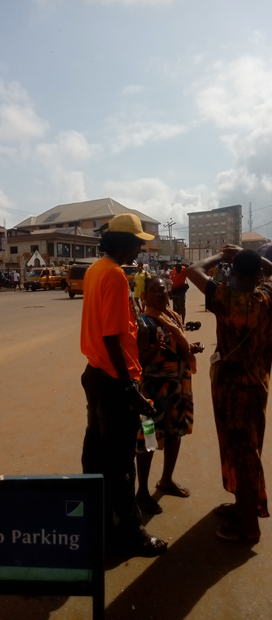 Three people stand together in an urban area of Nkpor, Nigeria. A man in an orange shirt and a yellow cap holds a bottle of Sprite. A woman in a patterned dress stands between him and another person. The background includes buildings, utility poles, other people, and yellow vehicles. A "Parking" sign is visible in the foreground. The ground is a reddish-brown dirt or pavement.