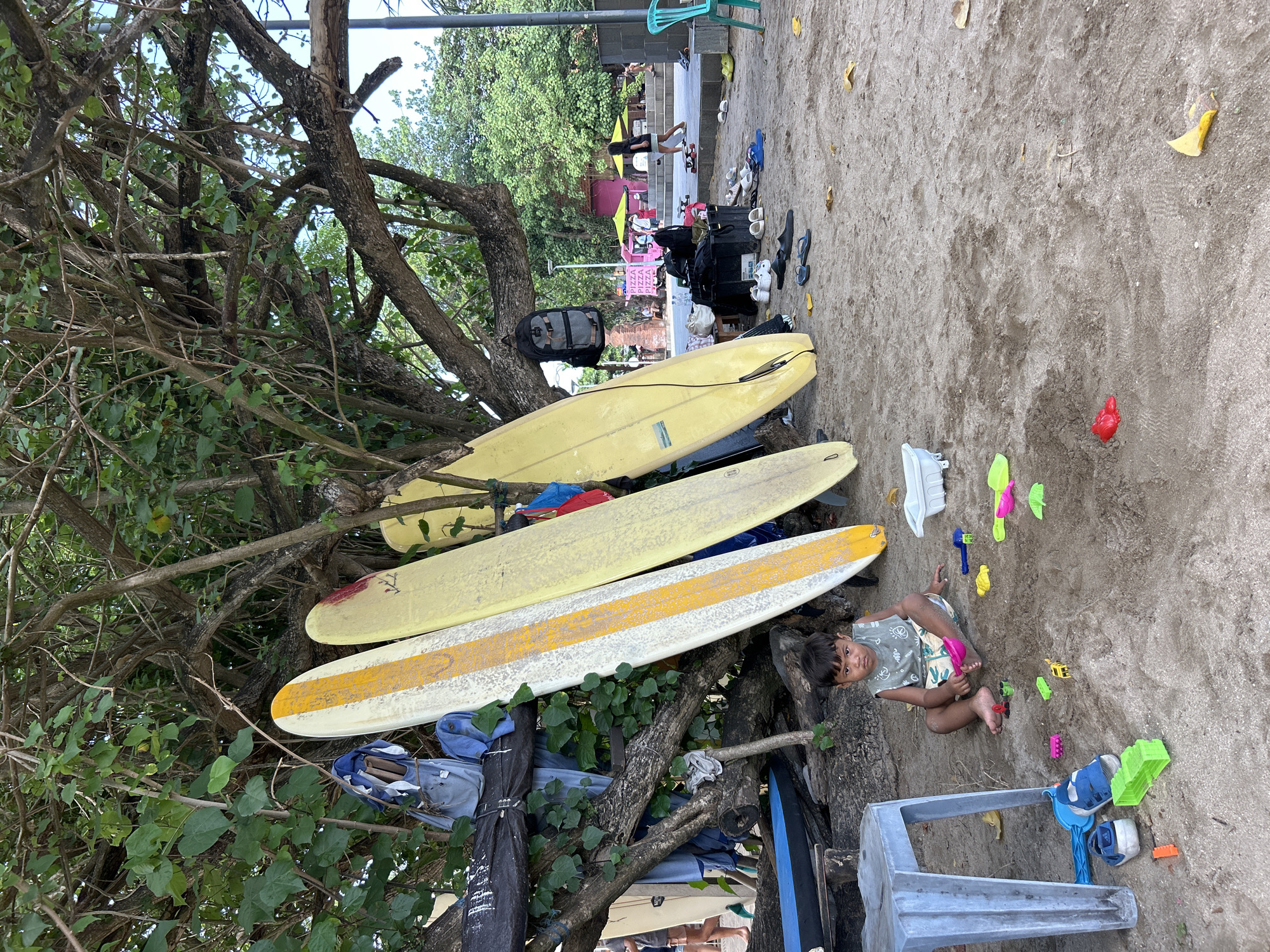 In Kuta, Indonesia, on a sandy beach beneath the shade of trees, a young boy sits on the sand, playing with colorful plastic toys, including shovels and molds. He is wearing a sleeveless shirt and shorts. Several surfboards lean against the trees behind him, some with distinct markings. A blue plastic chair is positioned to his left. In the background, people can be seen walking, and there are food stalls and more trees. The ground is covered in sand with scattered leaves and a white plastic bin. Several pairs of footwear are visible on the sand near the surfboards.