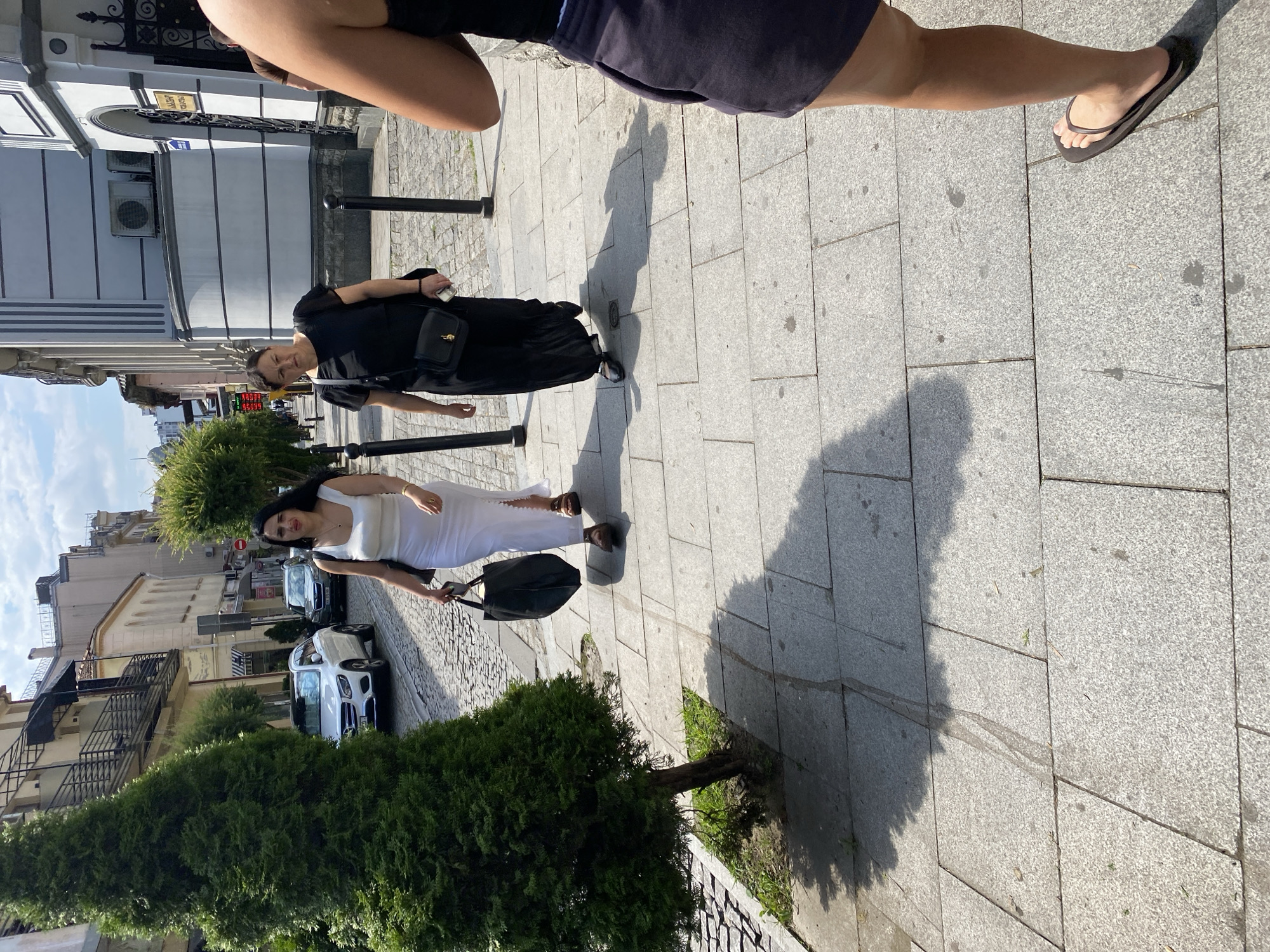The image depicts a sunny day on a cobblestone street in Batumi, Georgia, likely in the historic district, given the architecture. Two women are walking on a sidewalk. The woman on the left wears a white, sleeveless, floor-length dress and carries a large black tote bag. She is looking to her left with her mouth slightly open. The woman on the right wears a black, short-sleeved, floor-length dress and a black crossbody bag. She is looking forward and holding a smartphone in her right hand. A portion of a third person's legs, wearing a dark skirt and flip-flops, is visible in the foreground on the right. Trees and parked cars, including a white Mercedes-Benz, are visible on the street. The buildings have multiple stories with balconies and ornate architectural details. The ground is paved with stone slabs and cobblestones. Shadows from trees and people are cast across the sidewalk.