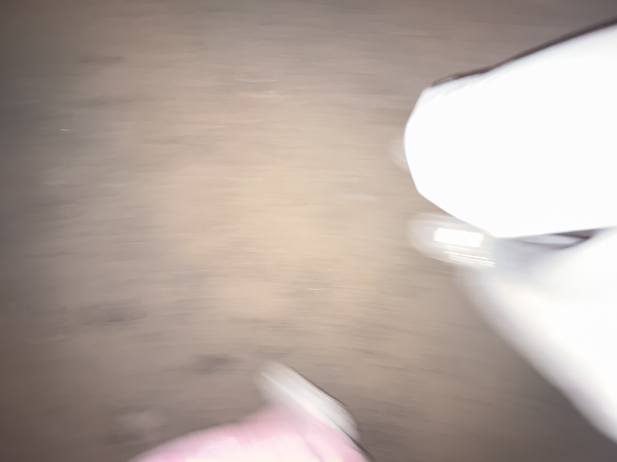 The image is a blurry, first-person perspective of someone walking on a brownish, textured surface. On the left side of the frame, a pink object, likely a boot or shoe, is visible. In the foreground, white pants and a white shoe are also visible, suggesting the person is wearing white trousers and white footwear. The blurriness of the image indicates movement. There is no discernible information about the location within the city of Dnipro, Ukraine, or any specific details about actions, interactions, or the environment beyond the immediate visual elements.