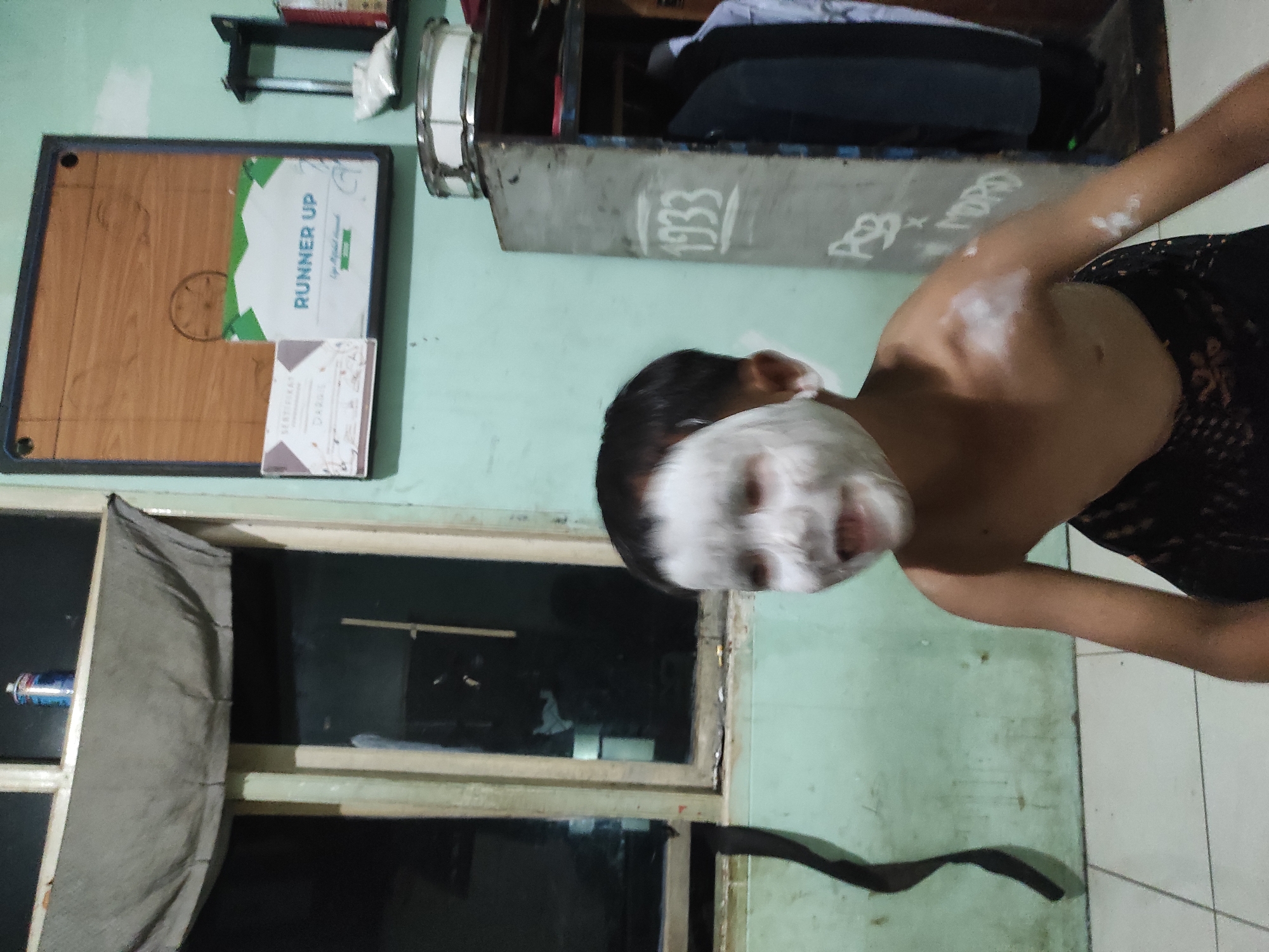 The image depicts a young person with white paste applied to their face and shoulders, possibly a mask. They are looking towards the camera with a wide smile and their mouth open. The person is shirtless and wearing a dark patterned sarong. The background appears to be an indoor setting with light green walls. A window with a grey curtain is visible on the left, and a cabinet with hanging clothes is on the right. A framed item with "RUNNER UP" text is mounted on the wall. The floor is tiled. The overall impression is of a casual, domestic environment. There is no indication of the image being located in Sumedang, Indonesia.