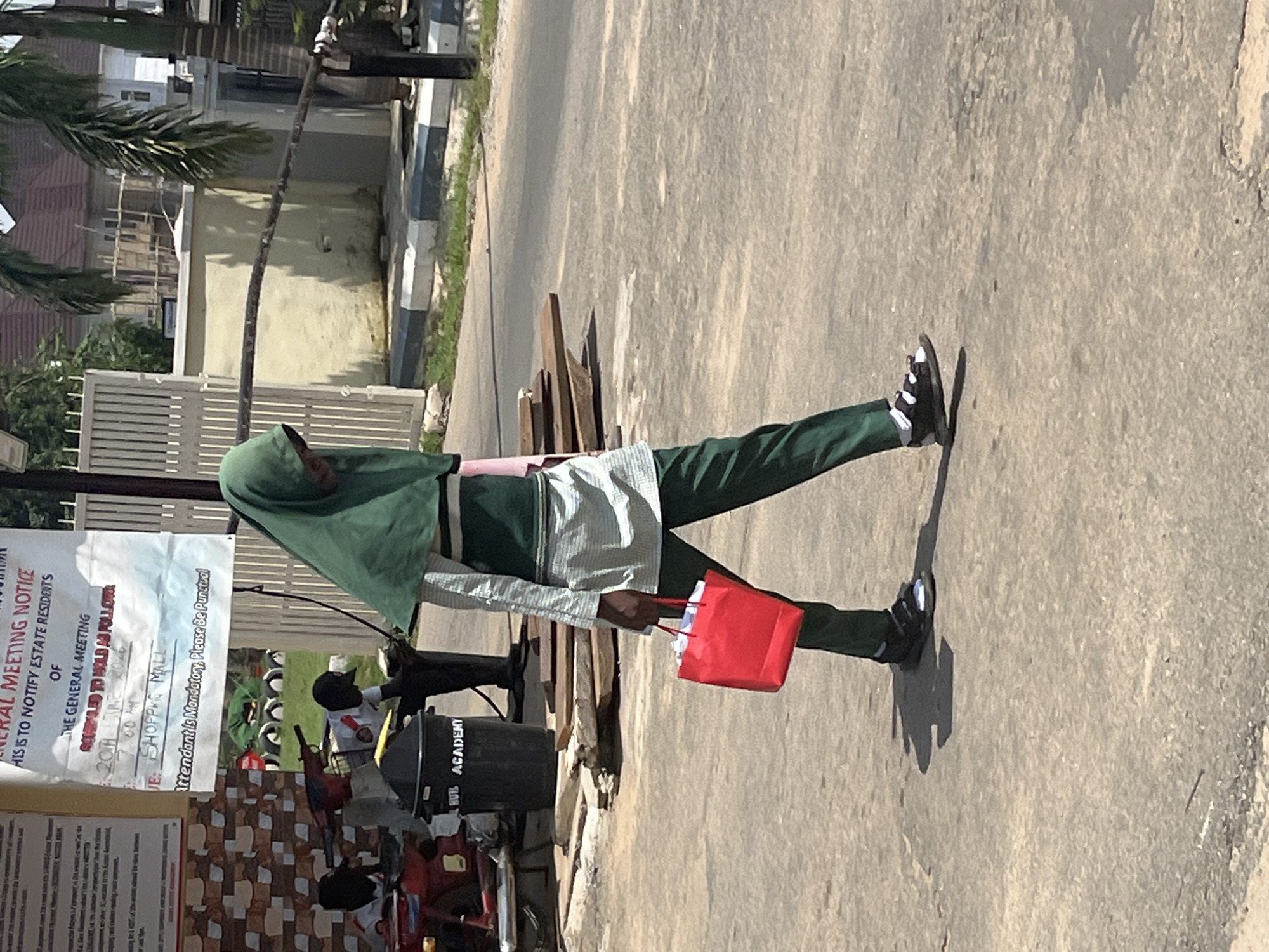 The media file shows a person, likely a student, wearing a green hijab and school uniform, standing on an asphalt road. They are holding a red shopping bag in their right hand. The person is dressed in a light-colored, long-sleeved shirt over a green uniform, with dark green pants and black and white sneakers. The image is taken outdoors on a sunny day, casting a distinct shadow.
Behind the person, a notice board with text is visible. The text on the notice board announces a "GENERAL MEETING" for "ESTATE RESIDENTS" to be held on "20TH JUNE 200..." at "7:00 AM" in "THE SHOPPING MALL". It also states that attendance is "Mandatory". Below this, "HUB ACADEMY" is printed.
In the background, there are residential buildings, palm trees, a fence, and some parked vehicles, including what appears to be a motorcycle. The scene suggests a residential area in Abuja, Nigeria. The lighting indicates it is daytime, likely morning given the meeting time mentioned on the notice. The overall atmosphere appears neutral, with no strong emotional cues evident from the subject.