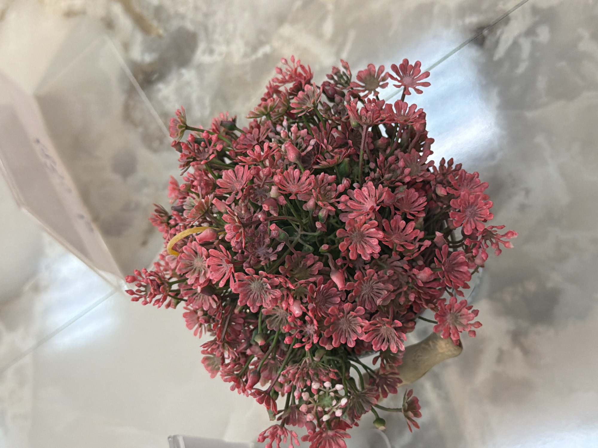 A close-up, high-angle shot shows a small artificial bouquet of pink flowers with green stems. The flowers have multiple small petals and appear to be clustered densely together. The bouquet is resting on a reflective, tiled surface that has a grey and white marbled pattern. In the background, out of focus, there is a clear plastic surface with some faint writing on it, and a white object to the left of the frame. The lighting suggests an indoor environment.