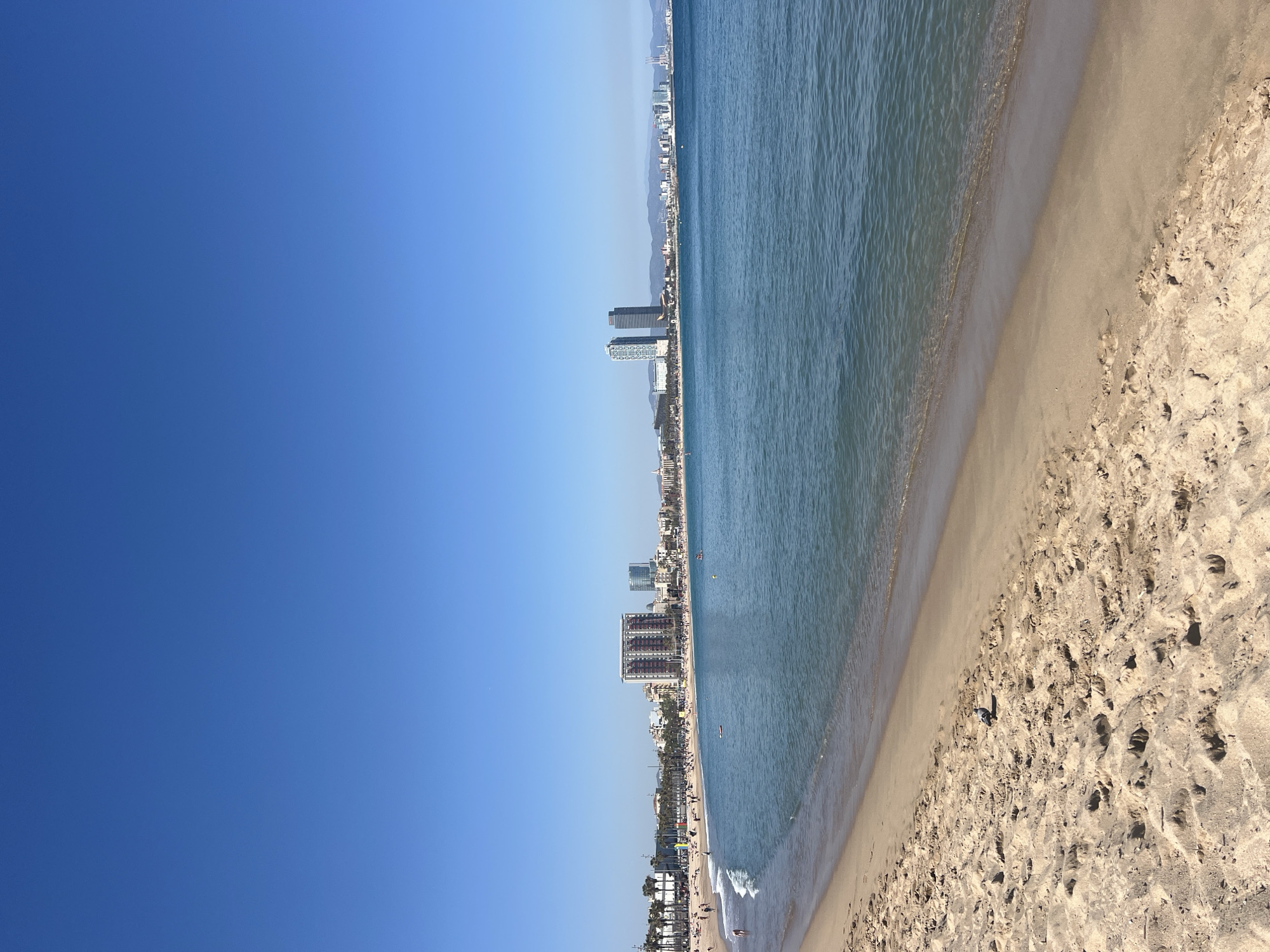 This is a beach scene in Barcelona, Spain, viewed from an elevated perspective. The image shows a stretch of sandy beach on the right, meeting the turquoise water of the Mediterranean Sea. Parallel to the beach is a city skyline, featuring a series of buildings that include tall hotels and apartments. The sky on the left is a clear, deep blue. The beach is populated with various beachgoers, and some people are seen swimming or wading in the water. The sand is marked with numerous footprints.