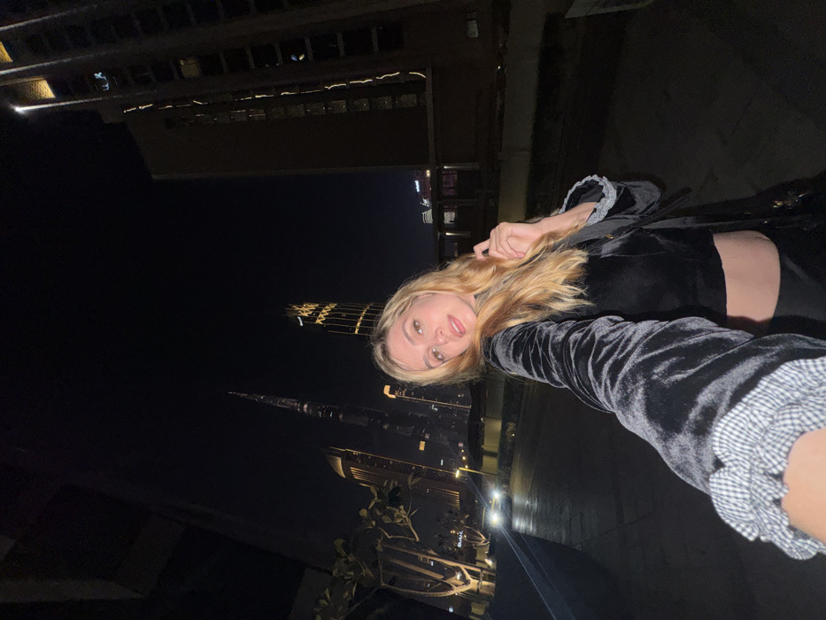 A blonde woman takes a selfie at night, with Dubai's skyline as the backdrop. The iconic Burj Khalifa is visible, lit up against the dark sky. The woman wears a black outfit and has blonde hair. Building silhouettes flank the skyline. The woman looks at the camera. The photograph is taken at an angle, causing the buildings to appear tilted.