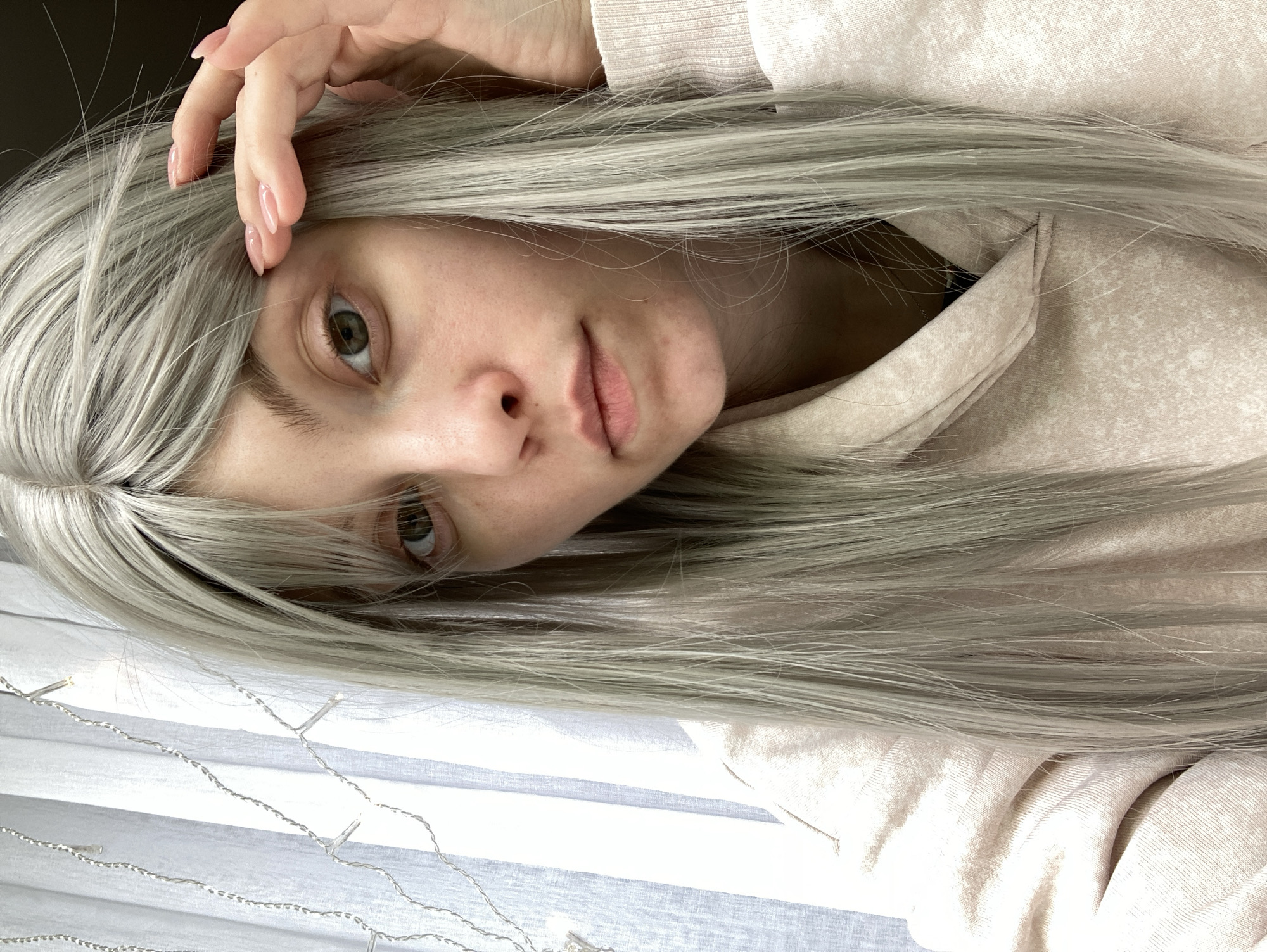 The image shows a close-up, selfie-style photograph of a young woman with long, light gray hair. She is wearing a light pink hooded sweatshirt. Her right hand is raised, with fingers touching her forehead and hair. She is looking directly at the camera with a neutral expression. The background consists of a white curtain and some string lights. There is no visible indication of the location being in Chaikovsky, Russia.