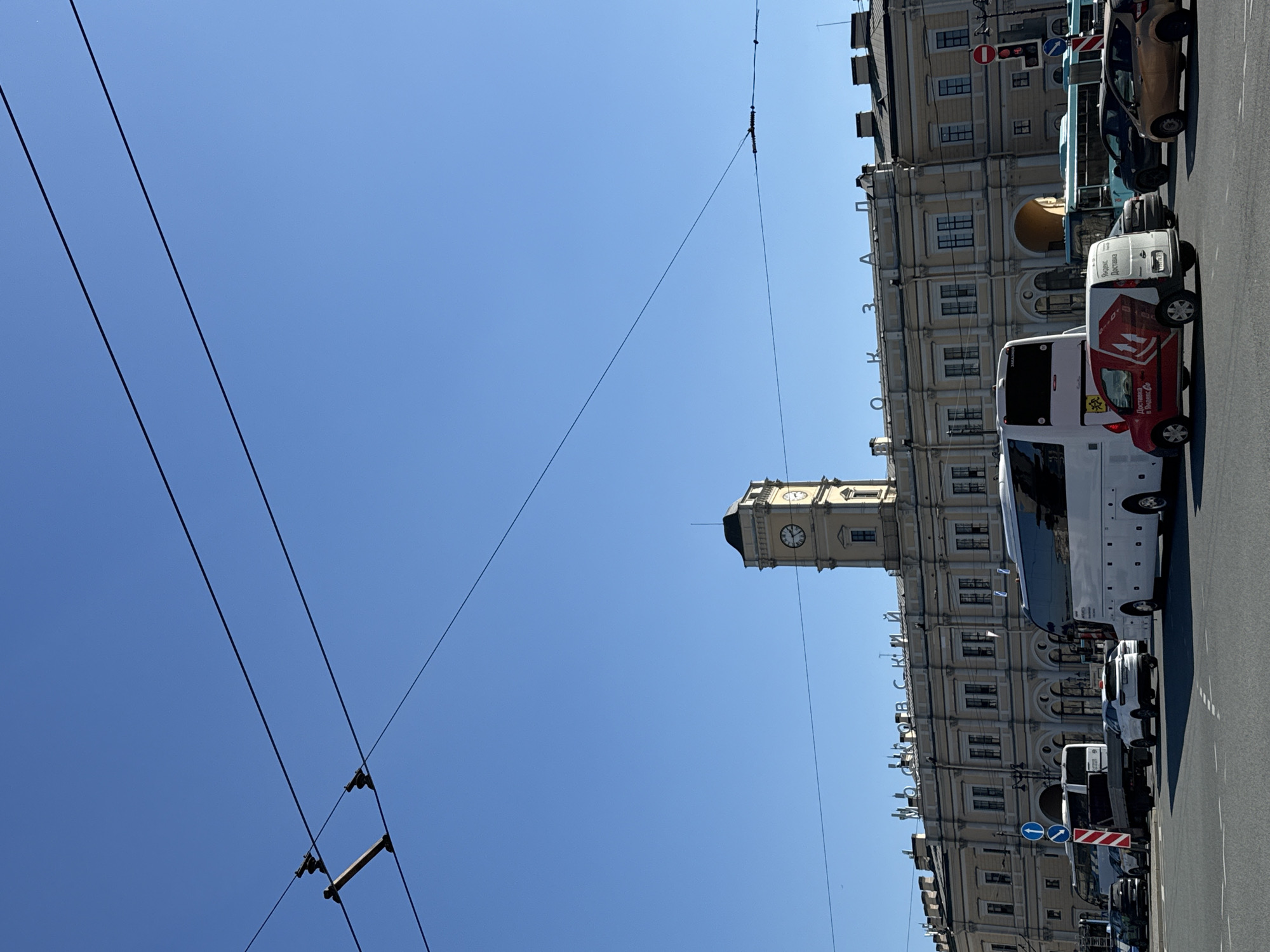 The image depicts a cityscape in Saint Petersburg, Russia. The sky is a clear, bright blue. Overhead electrical wires are visible diagonally across the upper left portion of the frame. In the right half of the image, there is a tall, light-colored building with a clock tower. The building is identifiable as the Moscow Railway Administration headquarters. Several vehicles, including buses and vans, are parked along the side of the street in front of the building. The overall composition is taken from a low angle, looking upwards towards the sky and building.