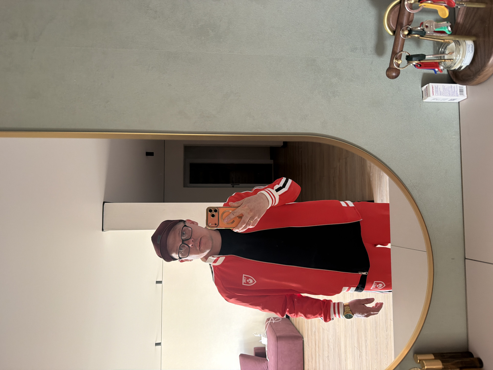 A man is taking a selfie in a mirror. The man is wearing a red jacket with white stripes on the sleeves, a black shirt, and a maroon beanie. He is also wearing glasses and a gold watch. The man is holding an orange iPhone. The mirror is oval-shaped and has a gold frame. The background is a modern interior with white walls and light wood flooring. There is a purple sofa in the background. There is also a key holder mounted to the wall. The image does not provide information that would identify the location as the city of Chaikovsky, Russia.