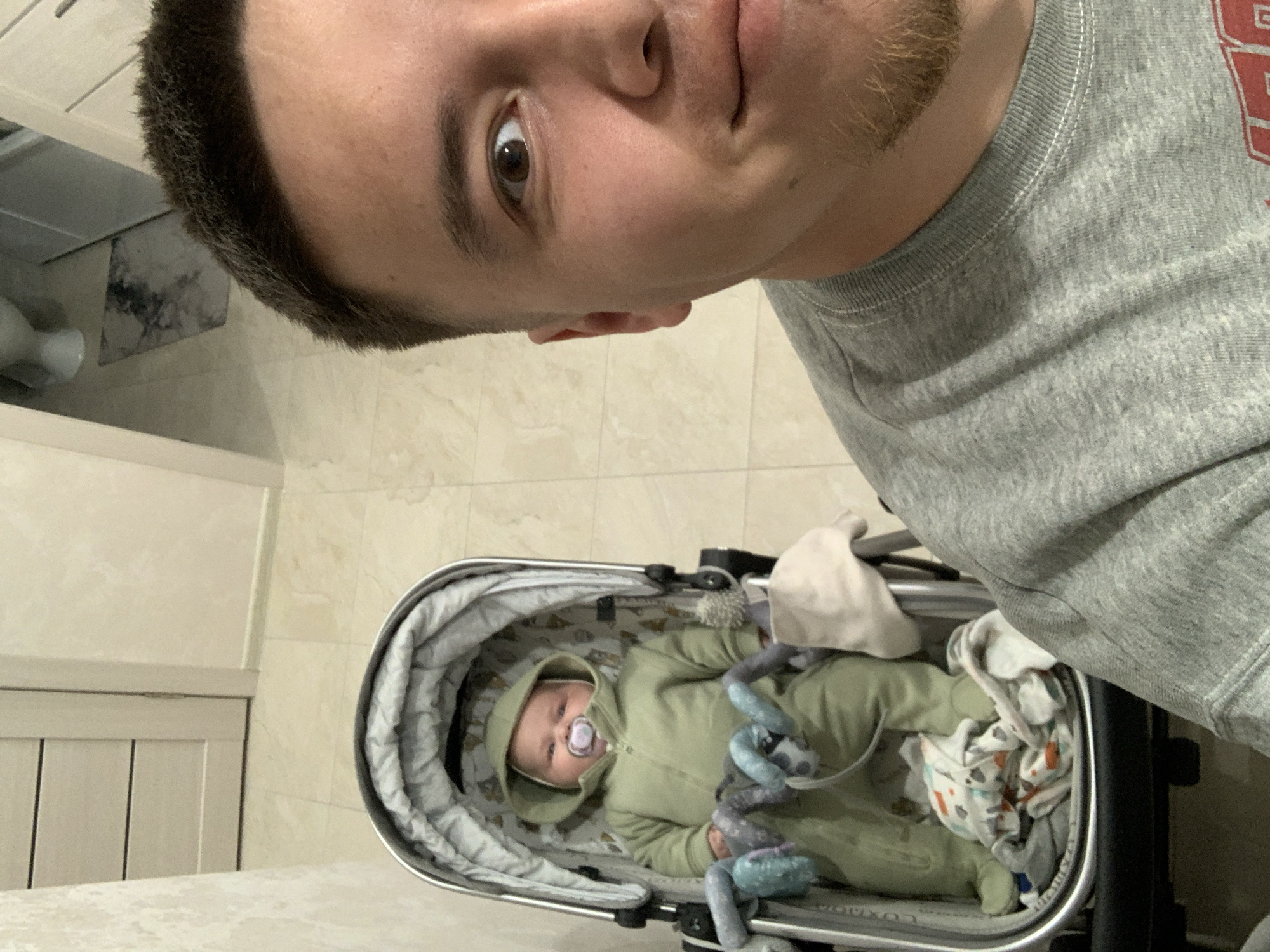 A close-up, selfie-style photograph shows a man and a baby in a stroller indoors. The man, positioned in the foreground on the right, is wearing a grey sweatshirt. His right eye is wide open and looking towards the camera, and he has a light beard. To the left of the man, and slightly further back, a baby is seated in a grey stroller. The baby is wearing a green hooded outfit and has a pacifier in its mouth. The stroller contains some blankets and toys. The background consists of tiled flooring and a portion of a closed door. The image was taken in Stavropol, Russia.