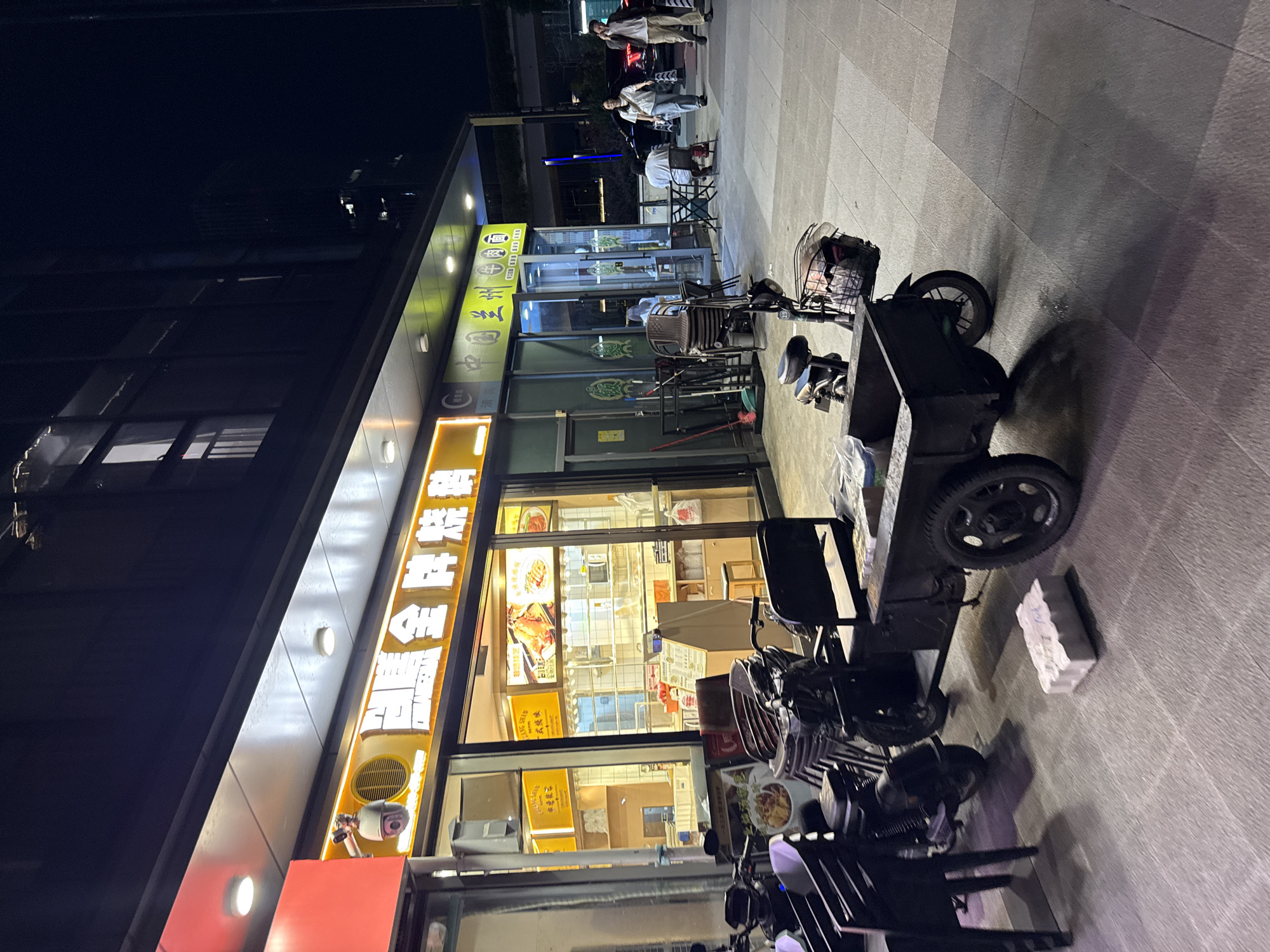 This image depicts a nighttime street scene in Tianhe District, China, focusing on the exterior of two adjacent food establishments. The left establishment features a bright yellow illuminated sign with Chinese characters, likely indicating a business name, and a security camera is visible above the entrance. The interior of this establishment is visible through a large glass window, showing food displays and kitchen equipment. In front of this store, several black plastic chairs and a motorcycle are parked.
To the right of this is another establishment with a green illuminated sign also bearing Chinese characters, identified as "China Lanzhou Beef Noodles." This eatery has glass doors and windows, revealing an indoor seating area where two individuals are seated at a table. Outside this second establishment, several chairs are stacked, and a red broom and dustpan are leaning against the wall.
In the foreground, a three-wheeled cart attached to a motorcycle is parked on the paved sidewalk. The cart appears to be for transporting goods. Several other motorcycles are also present. The ground is paved with large stone tiles, and shadows from the overhead structures and objects are cast across the scene. In the background, a dark building with illuminated windows and a glimpse of a cityscape are visible. Pedestrians, including two individuals walking and talking on cell phones, are present further down the street. The overall lighting suggests it is after sunset, with artificial lighting from the businesses illuminating the area.