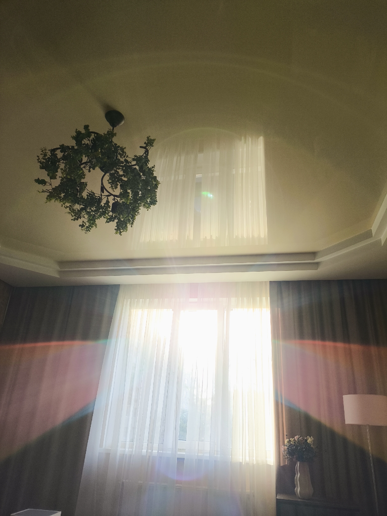 The image shows an interior view of a room with a large window covered by sheer curtains, allowing bright sunlight to stream in. Rainbow-colored light flares are visible across the image, particularly on the left side and on the ceiling. A chandelier with a leafy design hangs from the ceiling. To the right of the window, a floor lamp with a white shade and a vase of flowers on a surface are partially visible. The room appears to be furnished with dark curtains on the sides of the window. The ceiling has a tiered design. The overall impression is a brightly lit room with a focus on natural light and decorative elements. The scene is set in an unknown location, within an unknown city.