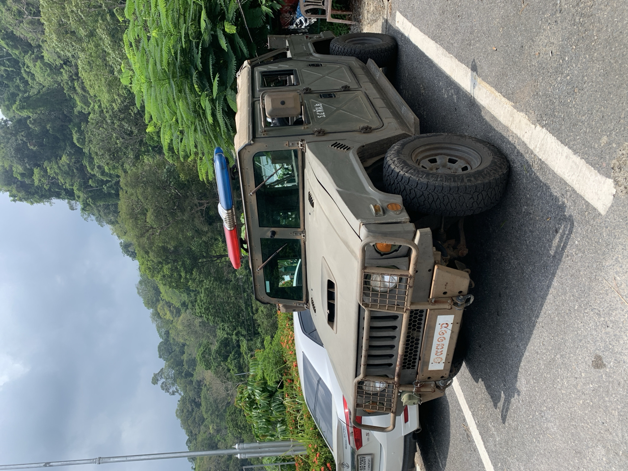 The image depicts a Hummer vehicle parked on a road next to a white car. The Hummer has a red and blue light bar on its roof and appears to be a military vehicle, with Thai writing visible on its front. The road has a white line marking the edge. The scene is adjacent to a lush green hillside and is within the city of Tambon Wang Prachan, Thailand.