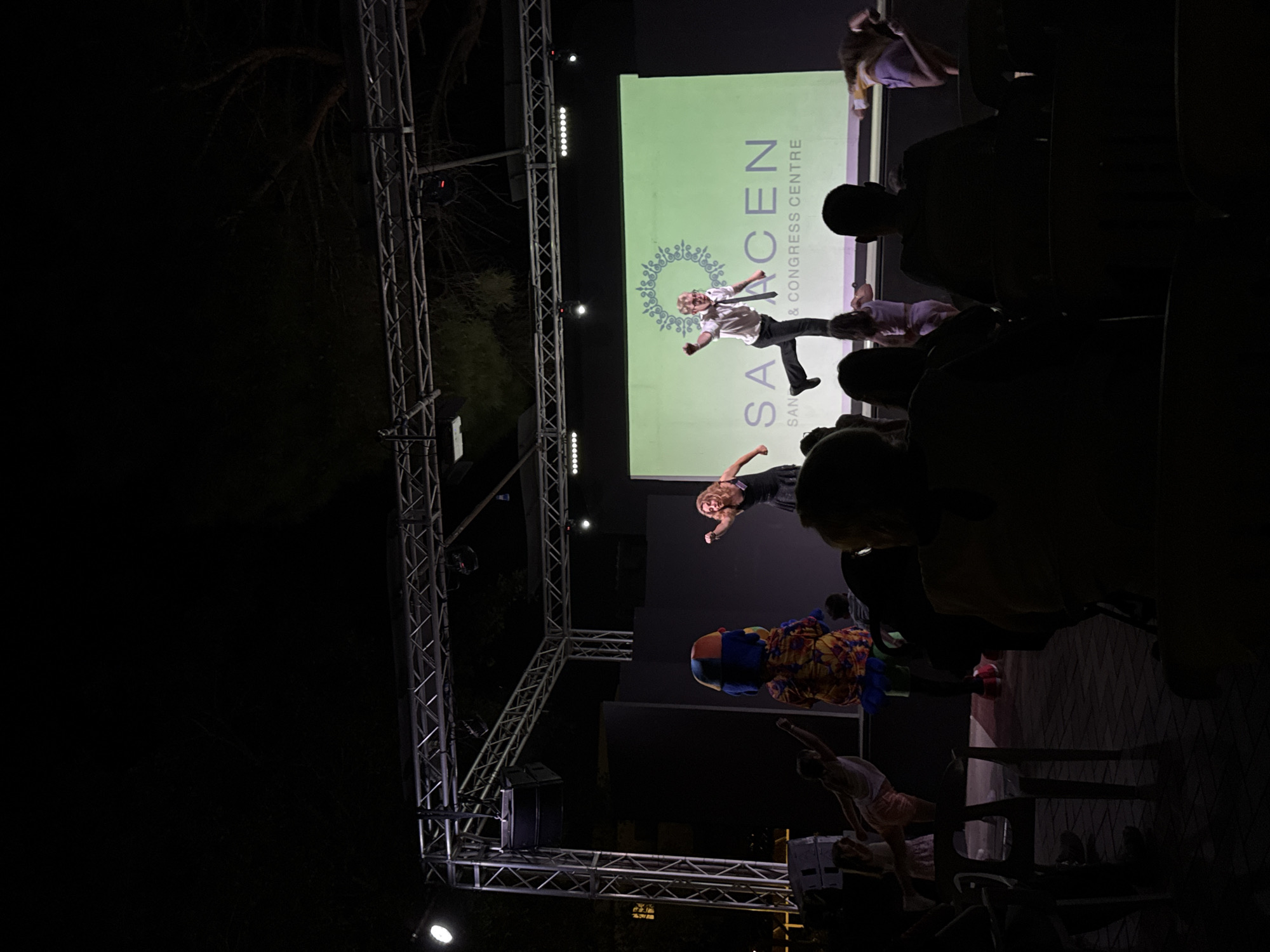 The image depicts an outdoor evening event at the Saracen San & Congress Centre in Isola delle Femmine, Italy. A stage with a large screen displaying the venue's name is set up, illuminated by spotlights and stage lighting. Two performers are actively engaged in a dance or energetic presentation on the stage. To the left, a person in a colorful costume, possibly a mascot, is interacting with younger performers. A crowd of spectators is seated in the foreground, silhouetted against the stage lights, suggesting they are watching the performance. The background shows trees and the dark night sky, indicating the performance is taking place outdoors.