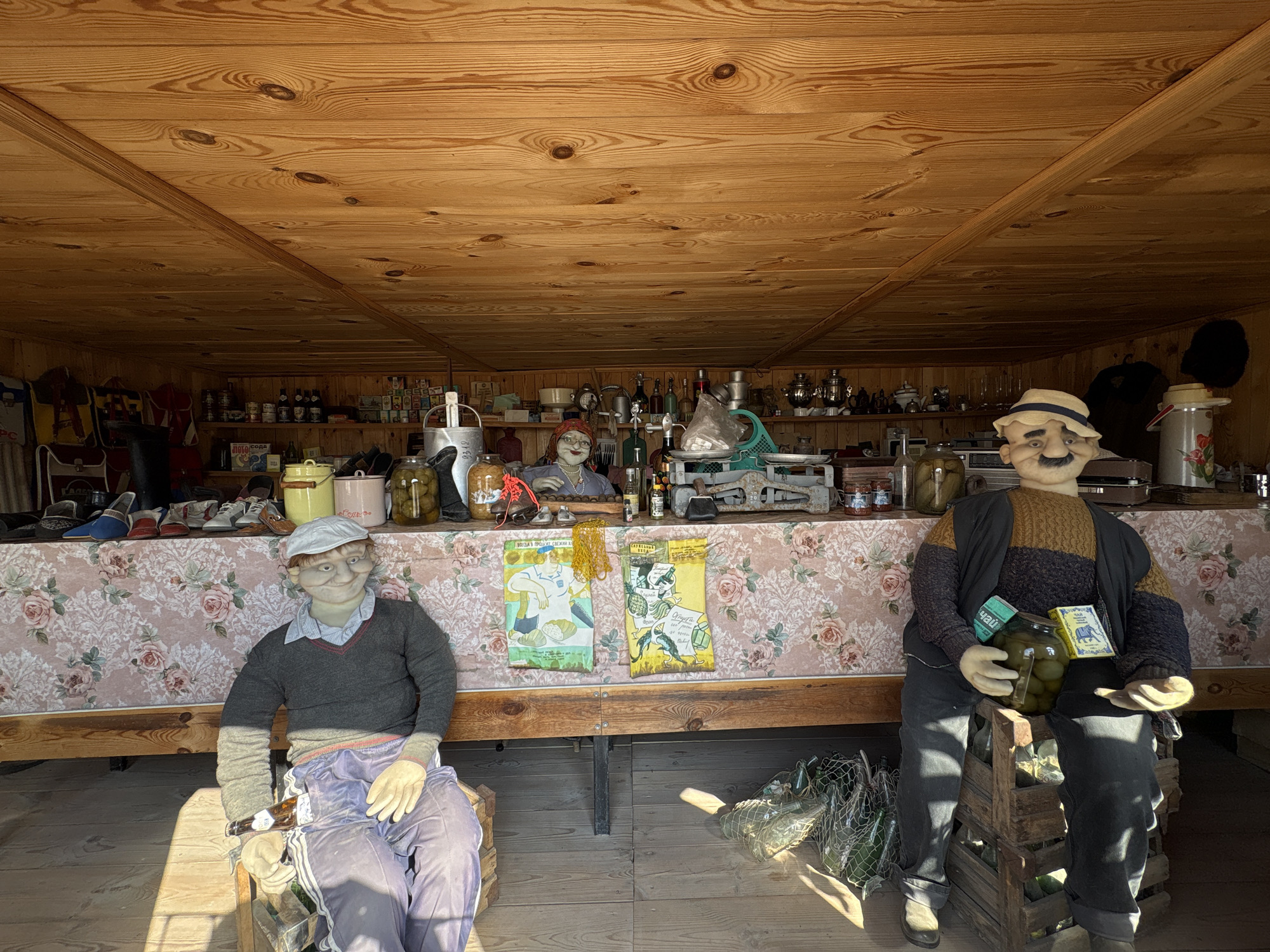 The image depicts the interior of a wooden structure in Bol'shoi Mashlyak, Russia. The space is lined with shelves filled with various items including bottles, jars, and decorative objects. Two mannequin figures are seated in the foreground, flanking a display of items on a table covered with a floral patterned tablecloth. The mannequin on the left is wearing a grey sweater and light purple pants, while the mannequin on the right is wearing a sweater and dark pants, and is holding a jar. Between them, two posters or leaflets are displayed. The space is illuminated by natural light.