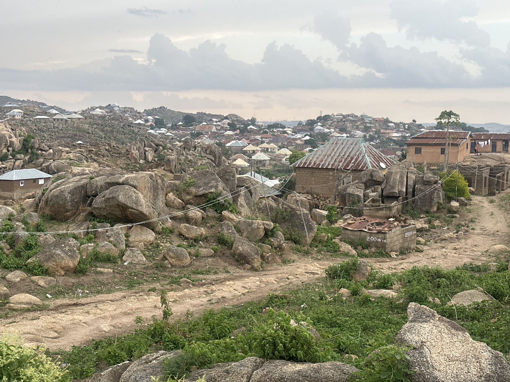 The image shows a rocky, hilly landscape with scattered houses in Jos, Nigeria. The foreground features large boulders and sparse vegetation. A dirt path meanders through the rocks and plants. In the midground, there are houses with metal roofs, some built among the rocks. In the background, more houses and hills can be seen under a cloudy sky.