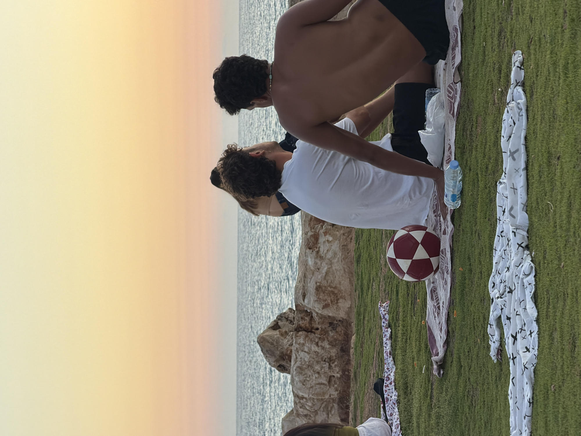 At sunset on a grassy area overlooking the Mediterranean Sea in Tel Aviv, Israel, two young men are seated on blankets. One man, wearing a white t-shirt, is facing away from the camera. The other man, shirtless and wearing dark shorts, is also facing away, with his back to the viewer. Beside them, a red and white patterned ball rests on a blanket. In the background, the sea stretches to the horizon under a gradient sky of orange and yellow. Large rocks line the shore. Several other people are visible in the background, seated and facing the sea. The overall scene depicts a relaxed gathering by the beach.