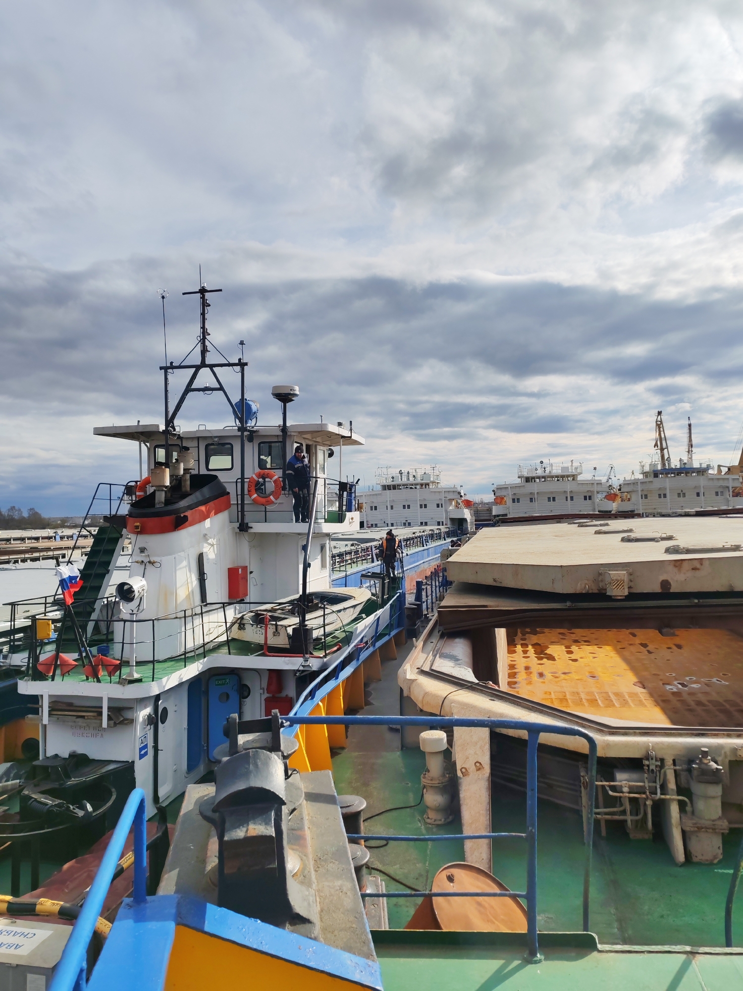 The image depicts a harbor scene in Rybinsk, Russia, under a cloudy sky. Multiple vessels are docked side-by-side. The foreground features the deck of one vessel, with blue and yellow painted railings, mooring equipment, and an orange barrel. A tugboat with a white and red superstructure is positioned to the left, displaying a Russian flag. Two figures are visible on the tugboat's upper deck. Adjacent to the foreground vessel is another, displaying a large rusty, rectangular container. Several white, multi-decked ships are visible in the background. A crane is also visible near the ships in the distance.