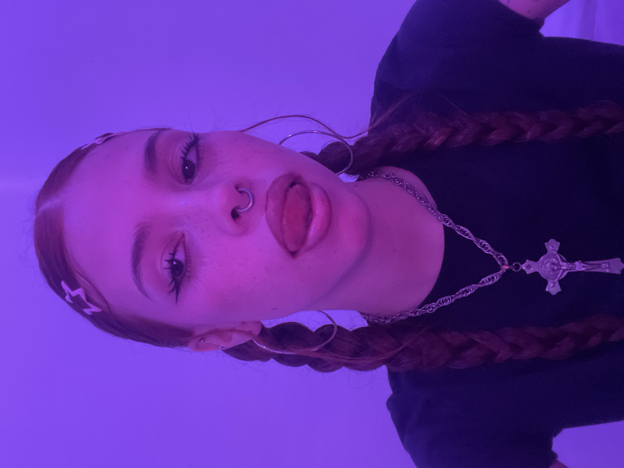 A young individual, likely female, with light skin and auburn hair styled in two braids, is positioned at a low angle, facing the camera. Her tongue is slightly protruded, and she is wearing a nose ring. She has hoop earrings and a silver chain necklace with a cross pendant. Her hair is adorned with star-shaped clips. She is wearing a dark-colored top. The background is a solid purple color. No elements indicate that the image was taken in Buenos Aires, Argentina, or any other specific location.