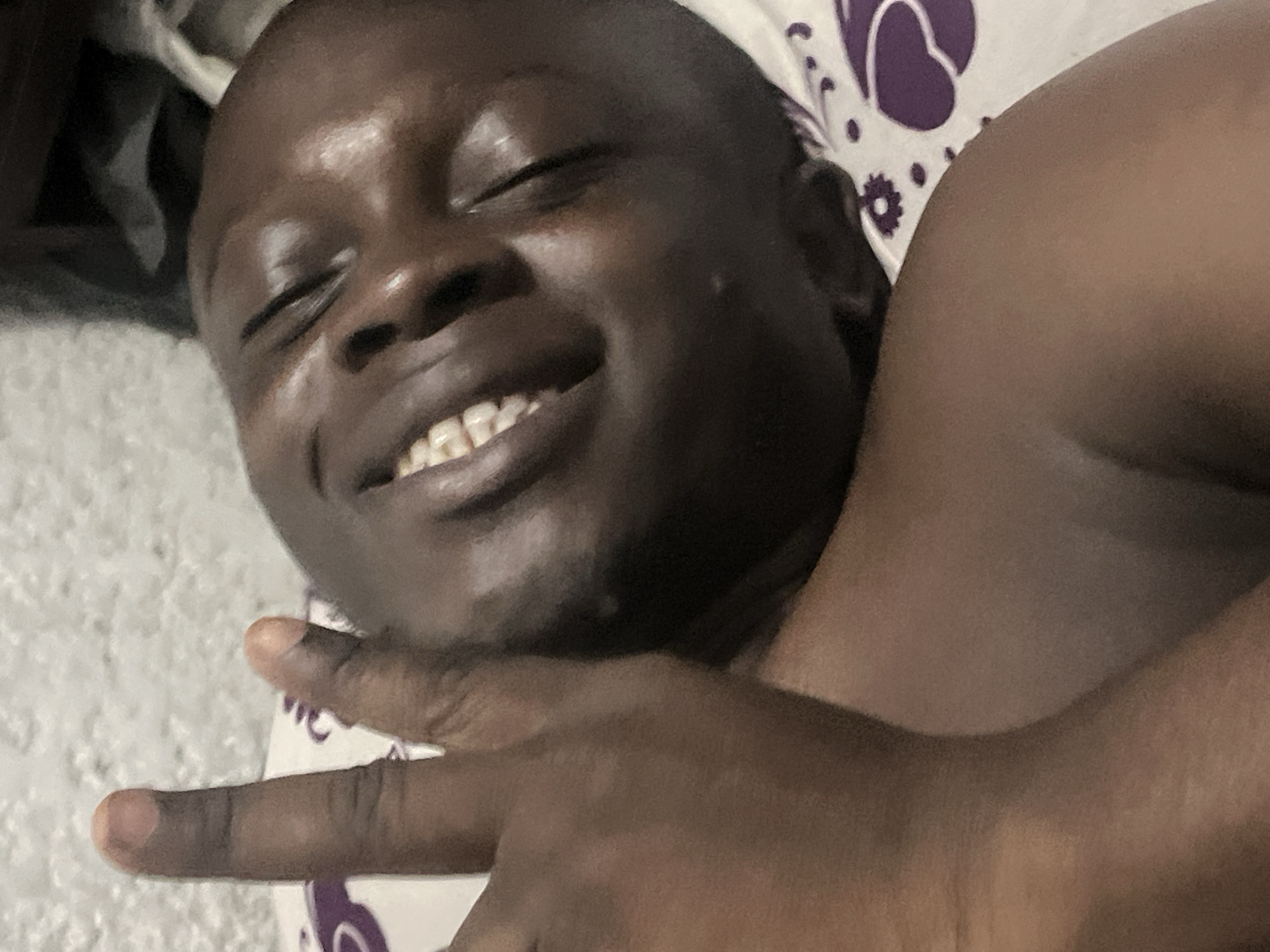 A close-up image depicts a smiling, dark-skinned person lying down. The person's eyes are closed, and their teeth are visible. A hand with fingers extended is positioned in the foreground. The person appears to be resting on a pillow or surface with a white and purple patterned design. The setting cannot be ascertained as Abuja, Nigeria based on the visual content of the image alone.
