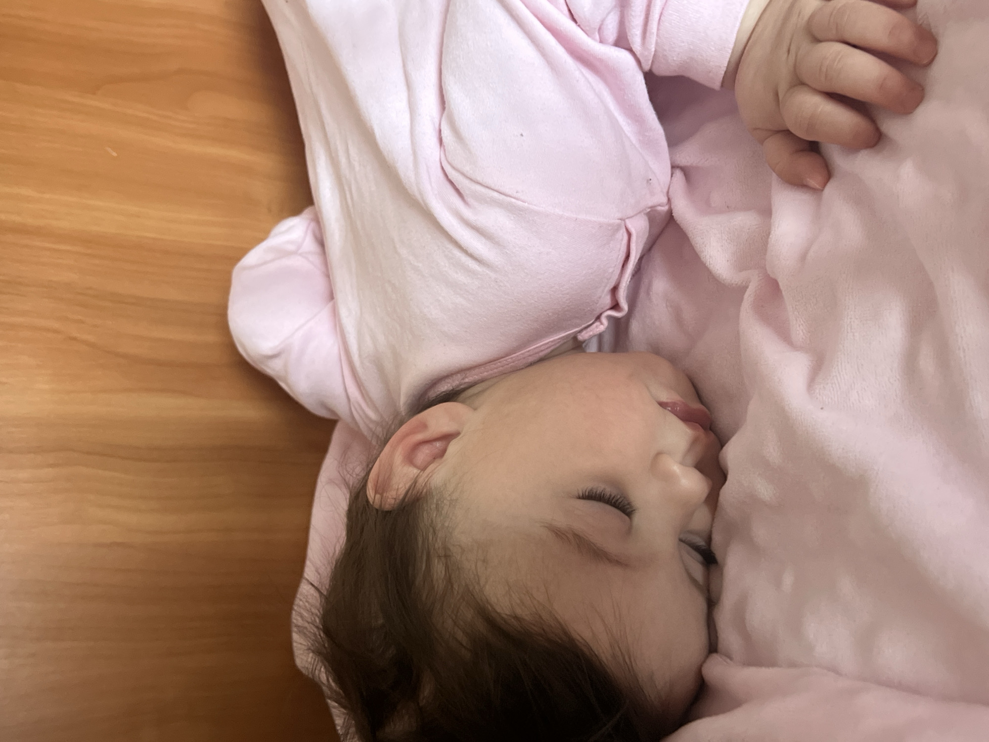 A baby is lying on its side on a pink blanket. The baby is wearing a pink outfit. The baby's eyes are closed, and its mouth is slightly open. The baby has dark hair and light skin. The blanket is wrinkled and soft. The background is a wooden surface. There is no indication of the location of Elena, Bulgaria in the image.