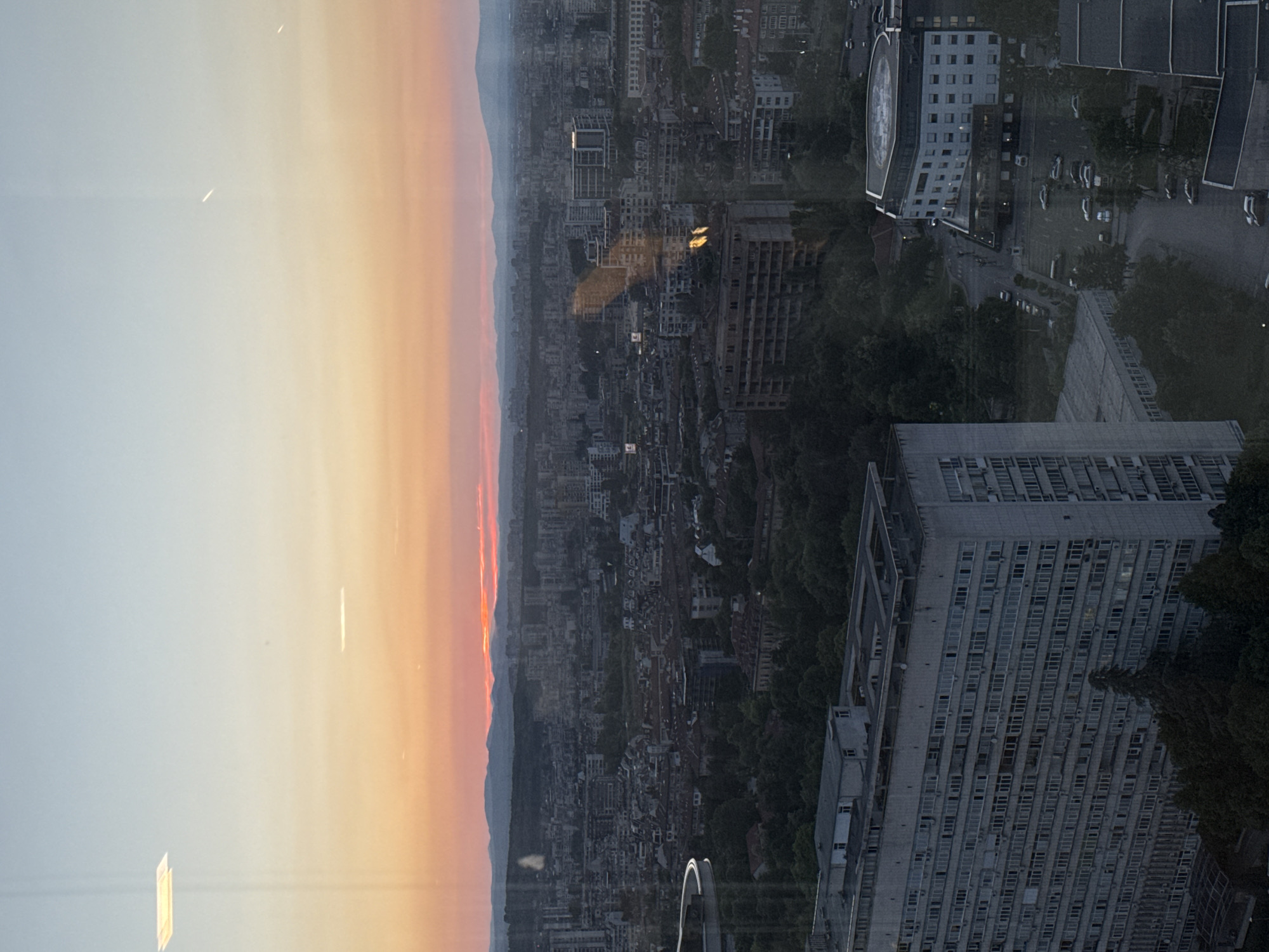 The image depicts a cityscape at sunset, possibly Sofia, Bulgaria, as indicated by the presence of a building resembling the NSA building on Aleksander Malinov Boulevard. The sky transitions from light blue to orange near the horizon, suggesting the time of day.
The city is densely built, with numerous residential and commercial buildings. Some buildings appear modern, while others seem older and possibly abandoned. There is greenery scattered throughout the urban landscape, with trees and parks visible between the buildings. The photo was taken from a high vantage point, likely from a tall building, overlooking the city. The reflective quality of the image suggests that it was taken through a window.