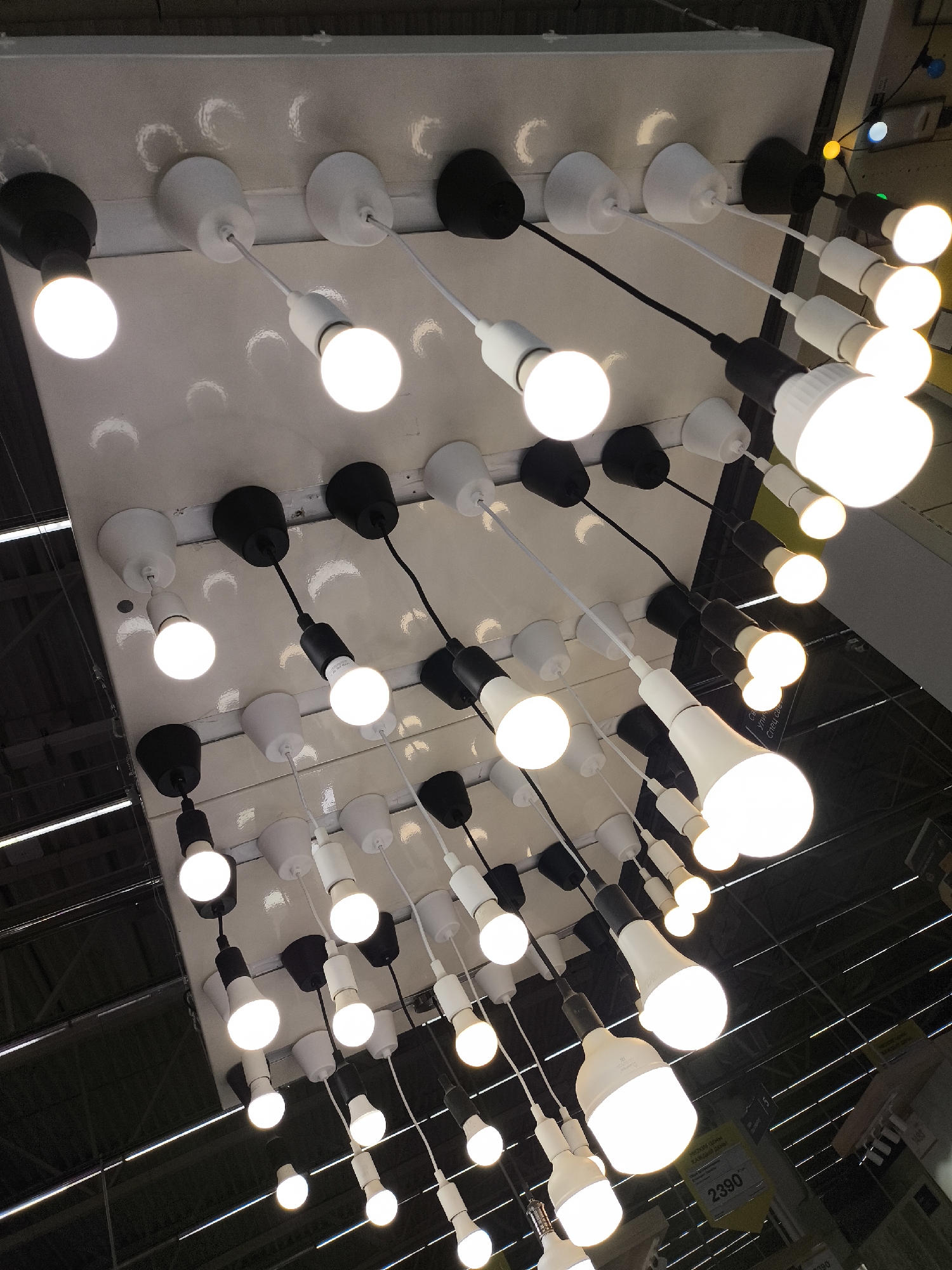 A low-angle shot depicts a display of numerous light bulbs hanging from a white rectangular ceiling fixture. The bulbs are of varying sizes and shapes, some spherical and others more elongated. Some are attached with black cords, others with white. The bulbs are arranged in rows and appear to be illuminated. The surrounding environment suggests a store or a showroom, as indicated by the ceiling structure and hanging lights, and a sign with the price 2390 is visible. While the location in Khimki, Russia cannot be verified from the image alone, the style of the store could be consistent with that region.
