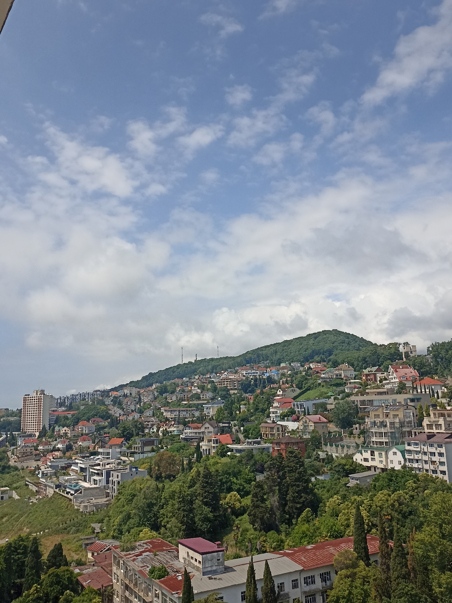 This is a panoramic, high-angle view of a densely populated hillside in Sochi, Russia, overlooking a bay. The city is built on steep slopes that lead down to the sea. Buildings range from modern apartment complexes and private residences with red-tiled roofs to older, partially-built structures. Lush green trees and vegetation cover the hillside between the buildings, creating a vibrant contrast with the urban development. The sky is partly cloudy, with white cumulus clouds scattered across a light blue expanse. Two tall communication towers are visible on the crest of the hill in the distance. The overall impression is one of a thriving coastal city nestled into a verdant, mountainous landscape.