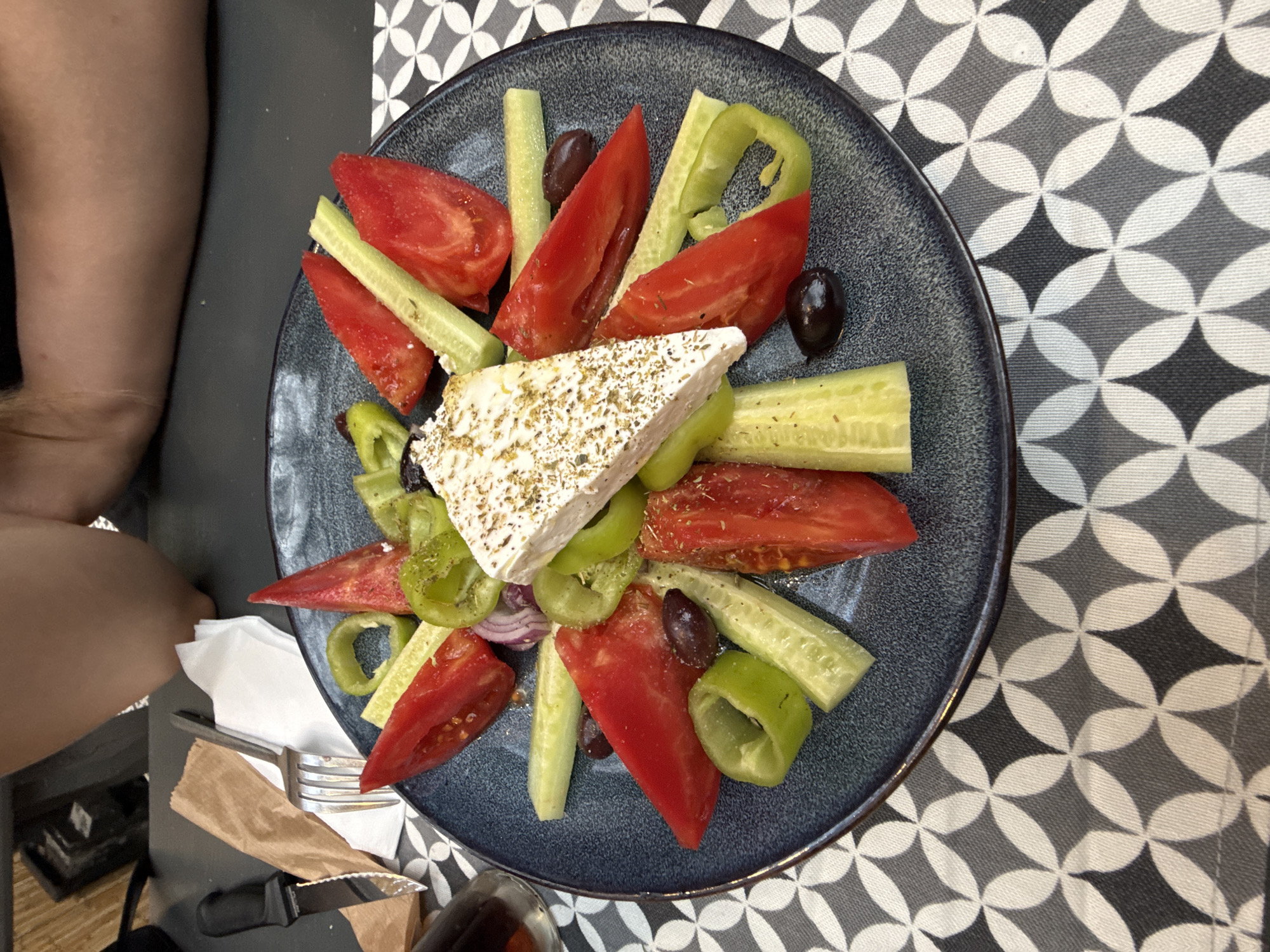 A close-up, high-angle shot shows a dark blue plate filled with a Greek salad. The salad contains a large wedge of feta cheese sprinkled with herbs in the center, surrounded by sliced red tomatoes, cucumber batons, green bell pepper rings, and Kalamata olives. The plate is set on a table with a grey and white patterned tablecloth. To the left of the plate, a knife with a black handle, a folded brown paper napkin, and a fork are visible. In the background, a person's lap and arms are partially visible, suggesting they are seated at the table. The scene is indoors, likely in a restaurant, in Stara Zagora, Bulgaria, given the information provided.