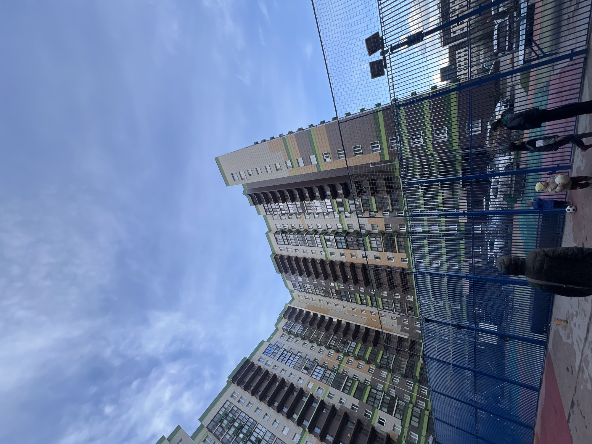 Captured from a low angle in Khimki, Russia, the image prominently features a high-rise apartment building with a staggered facade. The building exhibits a palette of beige, brown, and green accents along its edges and between windows, which are framed by dark balconies. To the right of the building, a blue metal fence and wire netting create a visual barrier. A few individuals are partially visible through the fence, and a soccer ball sits on the ground near them. The sky above is a mix of pale blue and white clouds.