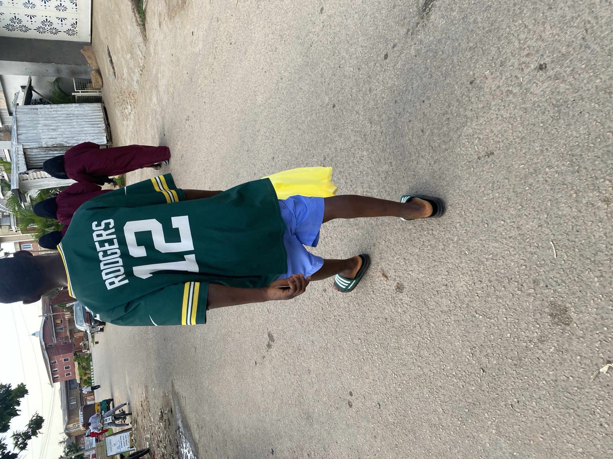A young Black person, seen from behind and slightly to the side, walks along a paved street. They are wearing a green jersey with yellow trim on the sleeves and collar, bearing the name "RODGERS" and the number "12" in white lettering on the back. Beneath the jersey, they wear light blue shorts and dark flip-flops with white stripes. A yellow piece of fabric is tucked into the back of their shorts.
The scene appears to be an urban street in Jos, Nigeria, with buildings and a dirt path visible in the background. Some of these buildings are made of corrugated metal. Other people are present in the background, including individuals wearing burgundy and dark clothing, one of whom appears to be wearing a hijab. A tricycle (Keke Napep) and a car are also visible on the street.
The lighting suggests it is daytime, likely late morning or early afternoon, with bright sunlight casting shadows. The overall mood is casual and everyday, with no specific event or strong emotions evident. The image seems to capture a moment of ordinary life on a street.