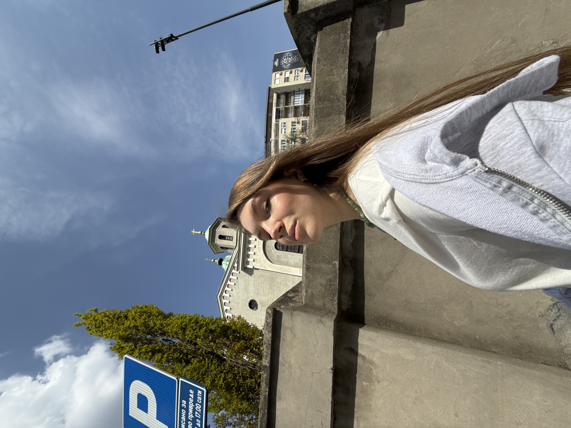The image, originally rotated 90 degrees counter-clockwise, captures a young woman outdoors in an urban setting on a sunny day. In the foreground, on the right side of the frame, a woman with long, straight light brown hair is visible from the chest up. She is dressed in a light-colored top and a grey zipped hoodie. Her face is shown in profile, looking slightly downwards and to the left with a neutral, contemplative expression. Sunlight brightly illuminates her hair and the side of her face.
Behind her, a concrete wall or barrier runs diagonally across the mid-ground. To the left of this barrier and in the background, an urban panorama unfolds under a clear blue sky dotted with scattered white clouds. A light-colored Orthodox church with green domes and golden crosses is prominently featured. Adjacent to the church, a lush green tree provides natural greenery. Further in the background, a taller, multi-story building with multiple windows and a dark billboard on its roof is visible. A street light or utility pole extends into the upper portion of the frame.
In the bottom left corner, a blue parking sign with a white 'P' symbol is clearly visible. The Cyrillic text on the sign reads: "ПАРКИРАЊЕ ЗА ВОЗИЛА ПРИВРЕДЕ ДО 17.00 САТИ", which translates to "PARKING FOR BUSINESS VEHICLES UNTIL 17:00 (5 PM)". The presence of this Cyrillic text and the distinctive Orthodox church architecture are strong indicators of a location such as Belgrade, Serbia, as suggested by the context. The scene is bathed in bright daylight, characteristic of midday or early afternoon, with strong illumination creating clear shadows. The overall impression is one of a calm, everyday moment in a bustling city.
