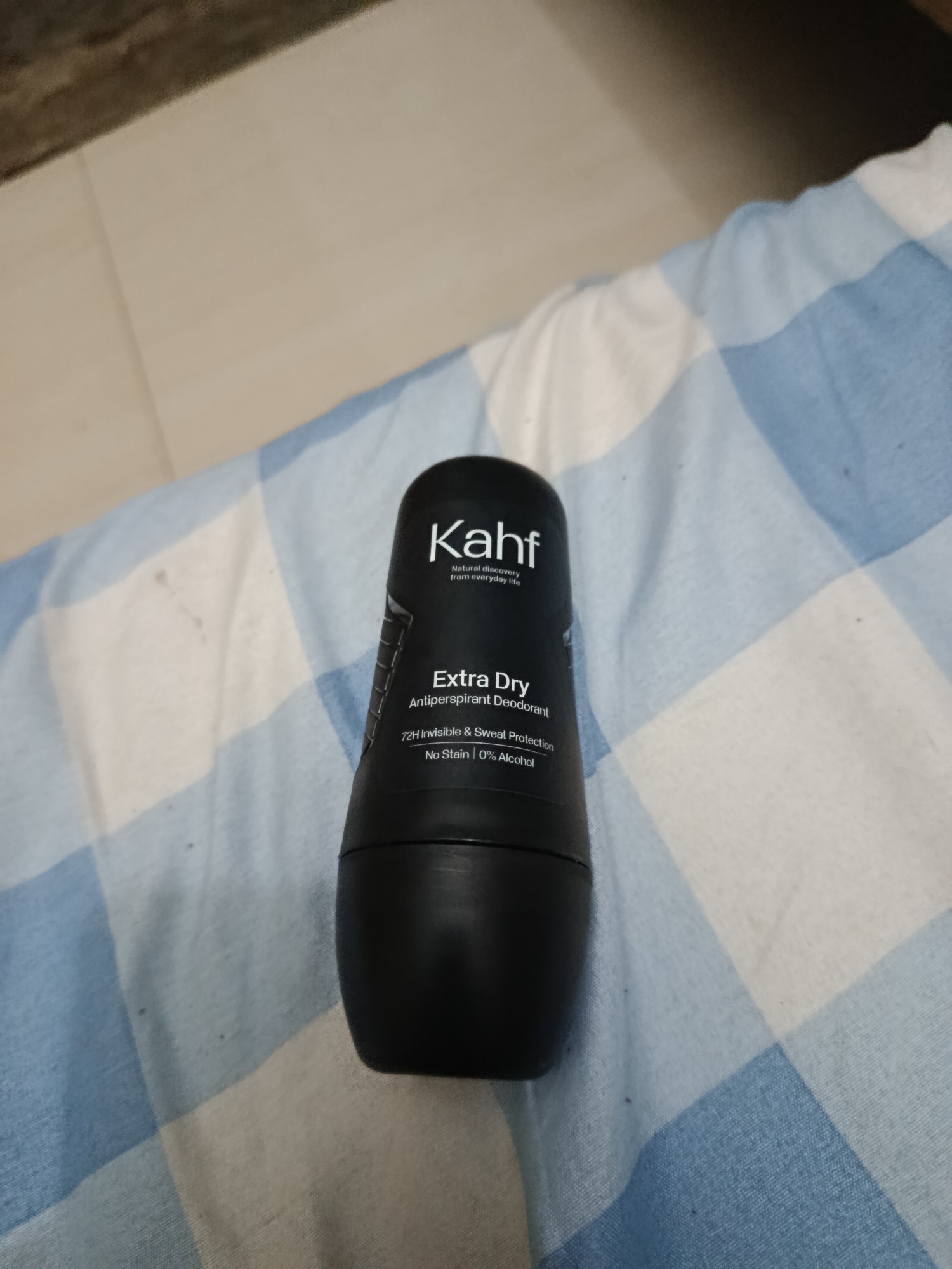 A black Kahf Extra Dry antiperspirant deodorant is placed on a blue and white checkered fabric. The deodorant has white text that reads "Kahf", "Natural discovery from everyday life", "Extra Dry", "Antiperspirant Deodorant", "72H Invisible & Sweat Protection", "No Stain | 0% Alcohol". The background is out of focus and appears to be a tiled floor with a dark upper section. This image was taken in Pacarmulyo, Indonesia.