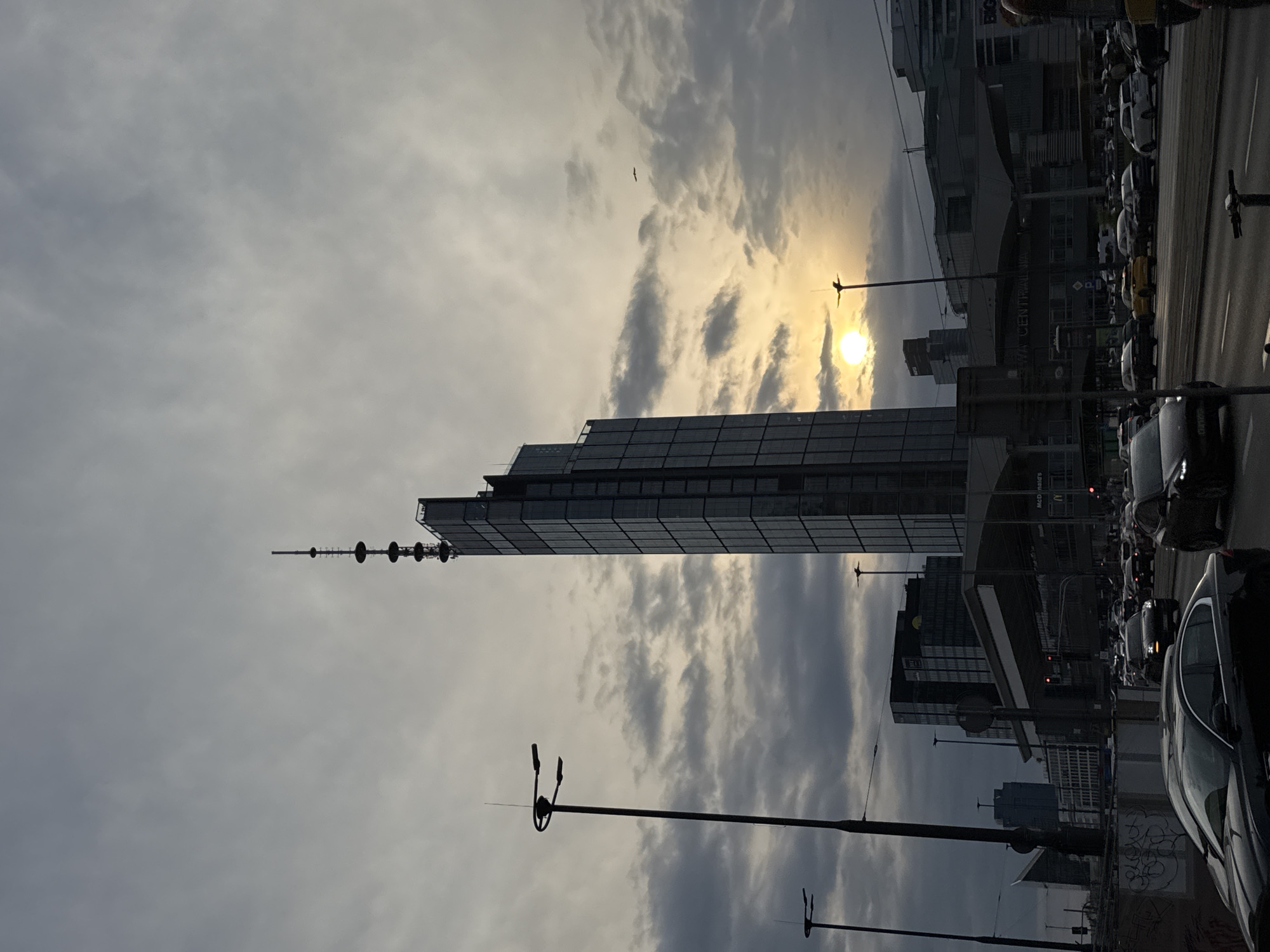 The image is a low-angle view of a city street in Warsaw, Poland, showing a tall skyscraper with a spire extending upwards. The sky is cloudy with the sun shining through the clouds. Street lights are visible in the foreground. On the right side of the image, there are several cars in traffic. The Warszawy Centralne train station is visible in the background.