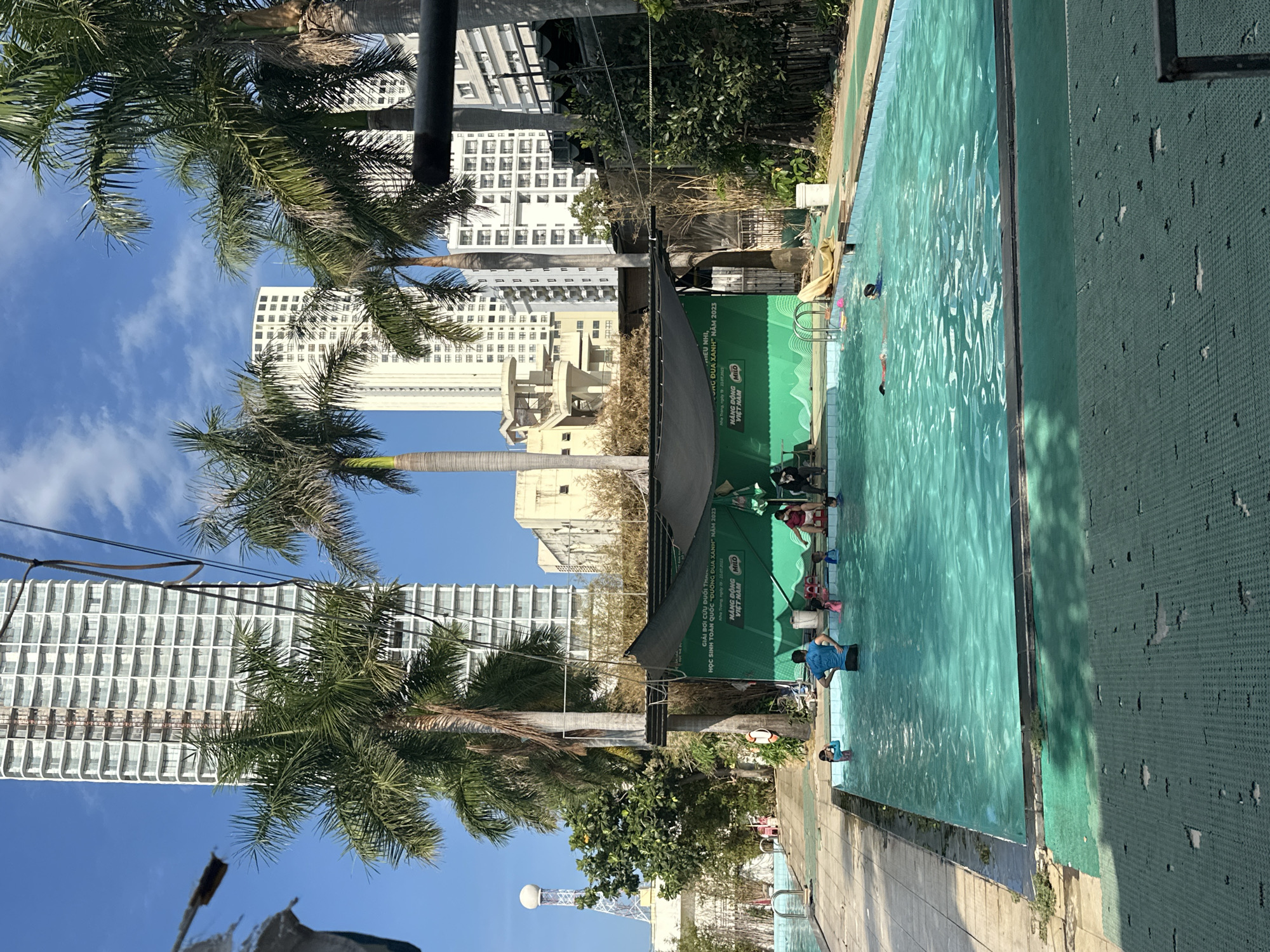The image captures a swimming pool scene in Nha Trang, Vietnam. The pool, bordered by cracked green tiles, reflects a light blue hue. A green banner with white lettering, reading "GIẢI BƠI CỨU ĐUỐI THANH," "HỌC SINH TOÀN QUỐC 'ĐƯỜNG ĐUA XANH' NĂM 2023," "Nha Trang, ngày 23.7.2023," "HANG BONG MILO," and "VIET NAM," flanks the pool, suggesting a swimming competition for students. Several people are in the pool, and some are standing along the edge, potentially supervising or coaching. Palm trees are visible in the foreground, and buildings rise in the background against a blue sky with scattered clouds. A communications tower is visible near the bottom of the frame.