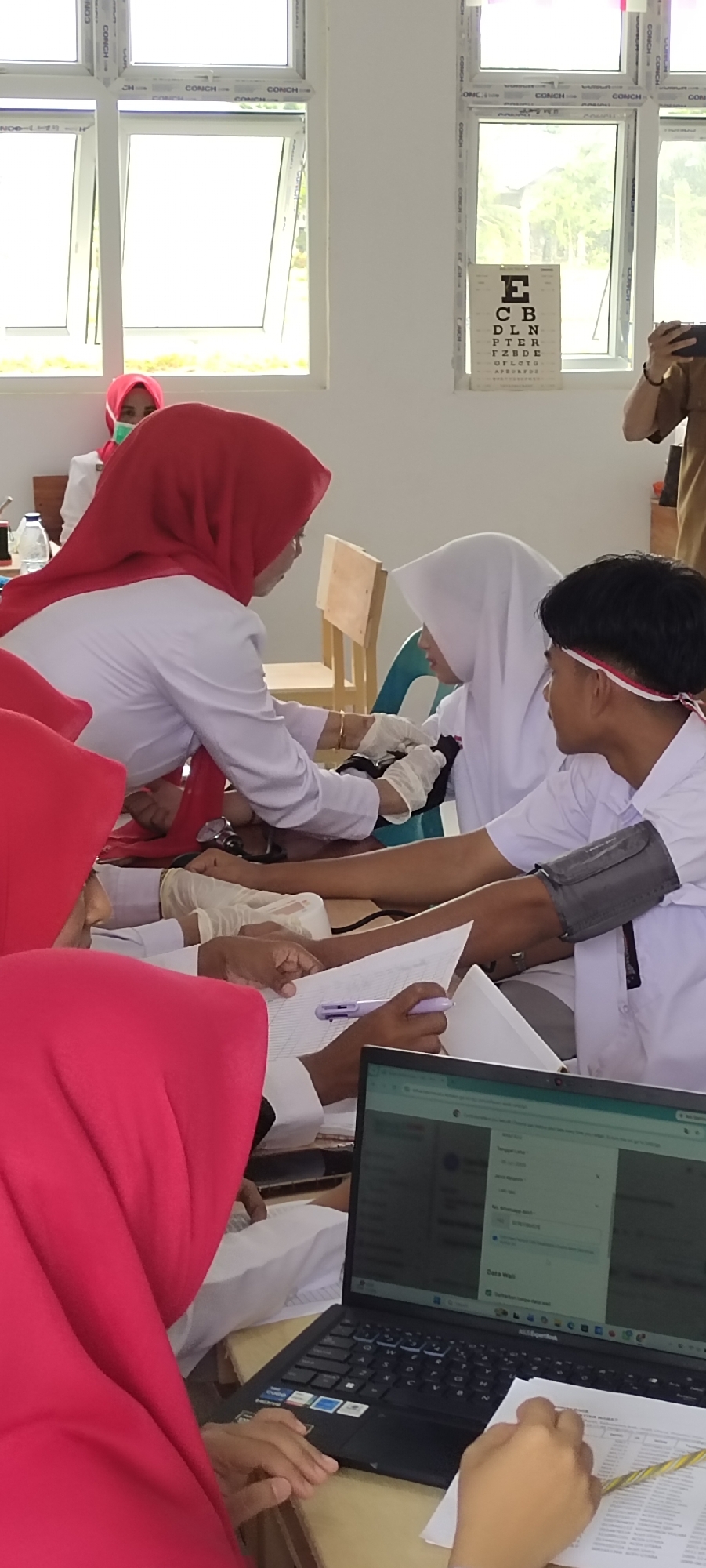In a brightly lit room with large windows, a medical examination is underway. Three individuals are actively involved in the scene. On the left, a person wearing a red hijab and white medical coat is carefully taking a blood pressure reading on another individual, who is also wearing a white hijab and a white medical coat, their arm extended for the cuff. In the background, another person in a red hijab and mask is seated, observing the procedure. To the right, a young man wearing a white shirt with a red and white headband is seated, and it appears someone is taking his blood pressure. A laptop is open on a table in the foreground, displaying what appears to be patient data or a medical application, with a pen resting on a medical chart. In the background, an eye chart is visible on the wall, suggesting the facility may also offer vision testing. The environment appears to be a clinic or a training facility.