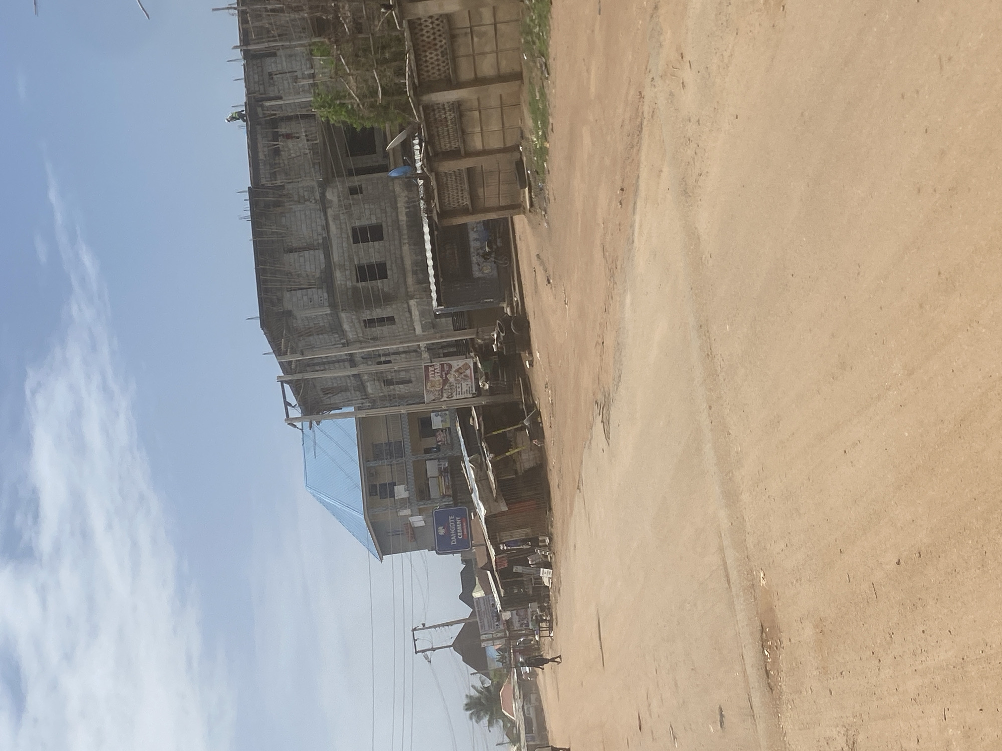 The image depicts a street scene in what appears to be a developing area. A multi-story building under construction dominates the left side of the frame. The building is made of grey cement blocks. Adjacent to it is a smaller building with a blue roof, indicating established commercial activity. Street-level businesses with signage are visible in front of the building, including a “DANCOTE CEMENT” sign. Power lines and poles run along the street. The road is unpaved and sandy. People are walking along the street. Based on the architecture, environment, and signage, the location is likely a commercial zone within Abuja, Nigeria.