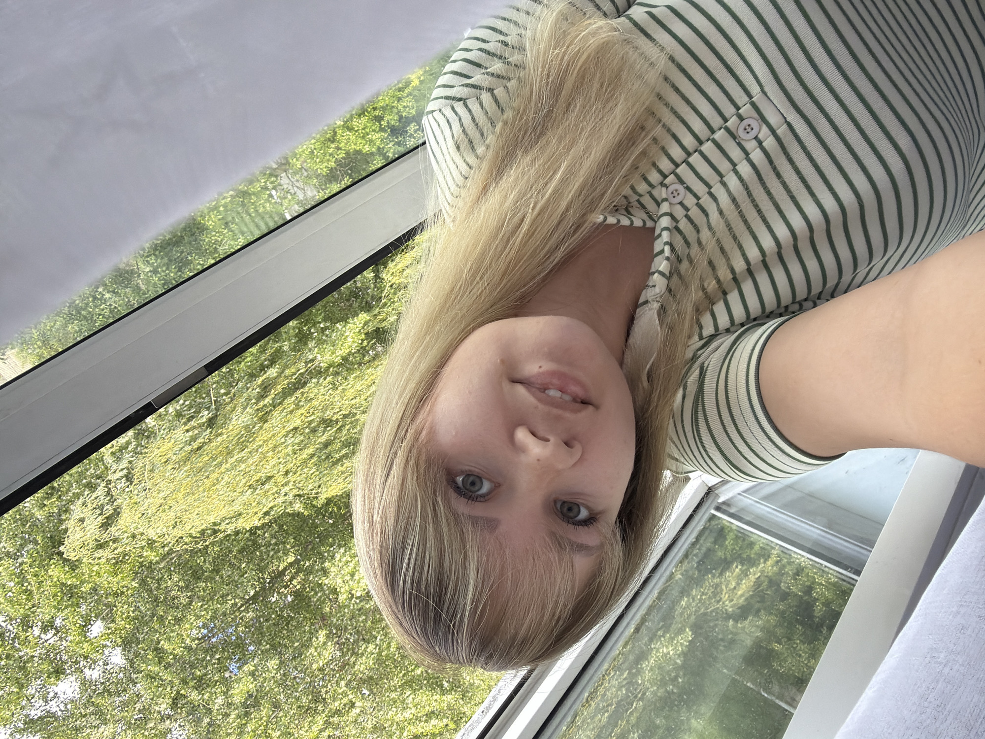 A young woman with blonde hair and blue eyes is taking a selfie indoors. She is wearing a striped, button-down shirt. The background shows a window with a view of green trees. While the photo's location in Chaikovsky, Russia is not discernible from the image, the window suggests a residential setting.