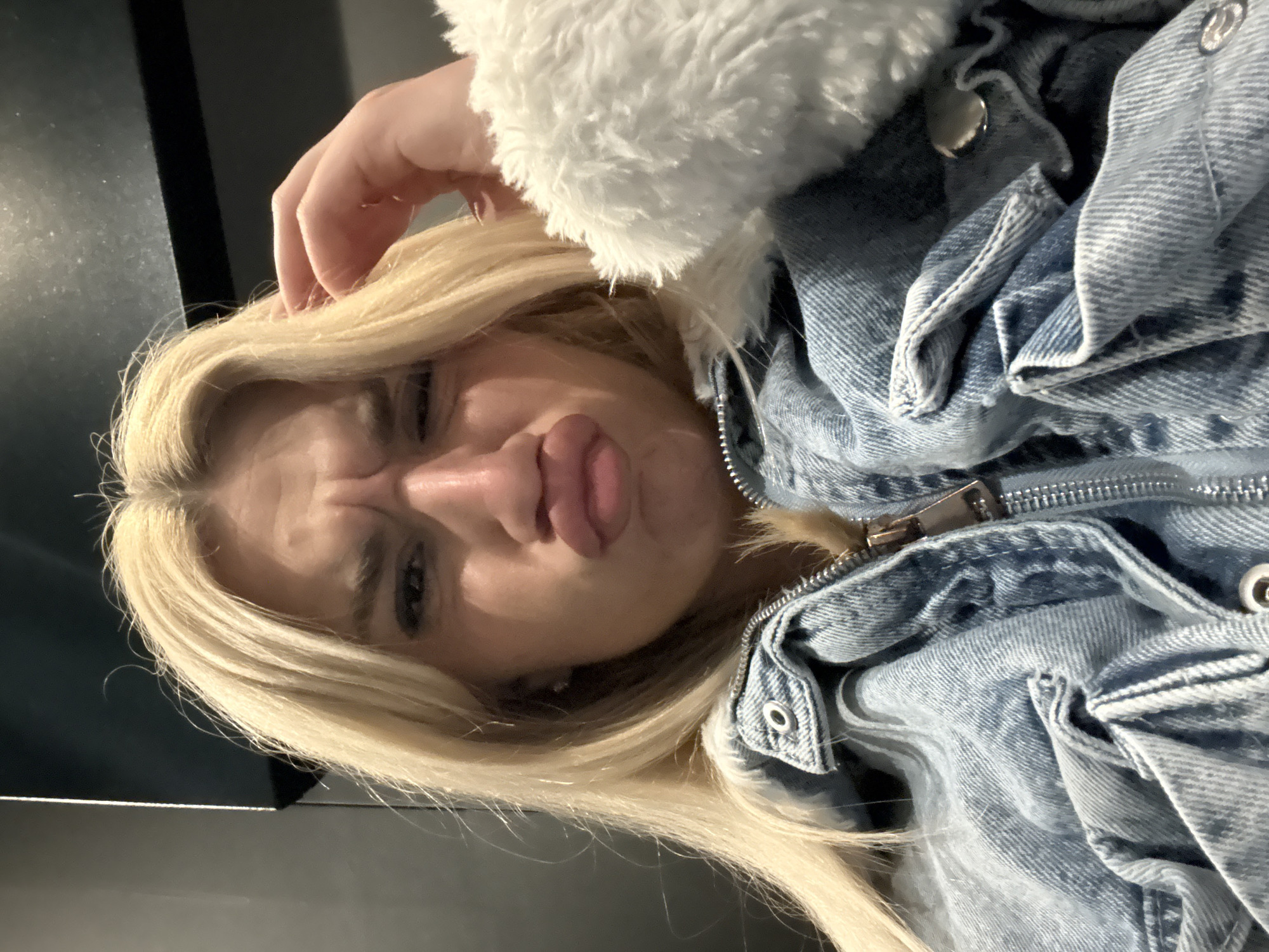 A close-up, angled selfie captures a young person with long, blonde hair, predominantly on the left side of the frame. Their head is tilted, giving the impression they are lying on their side. The subject is making a playful "duck face" or puckering their lips significantly, with their nose crinkled, indicating a humorous or exaggerated expression.
They are wearing a light blue denim jacket with a textured, fluffy white collar or inner lining visible around the neck and shoulder. The jacket features a prominent silver zipper down the front and multiple metal snaps or buttons on the denim fabric. One hand is partially visible at the top of the head, lightly touching the blonde hair.
The background on the left side of the image is dark and out of focus, appearing as a plain, unidentifiable surface, possibly a wall or door. A lighter, horizontal strip is visible at the very bottom left, suggesting a reflective surface like a table or counter. The lighting is artificial and close-up. No specific location cues are visible within the image to suggest Sofia, Bulgaria, though this is the provided context. There is no visible text. The overall mood conveyed is lighthearted and expressive.