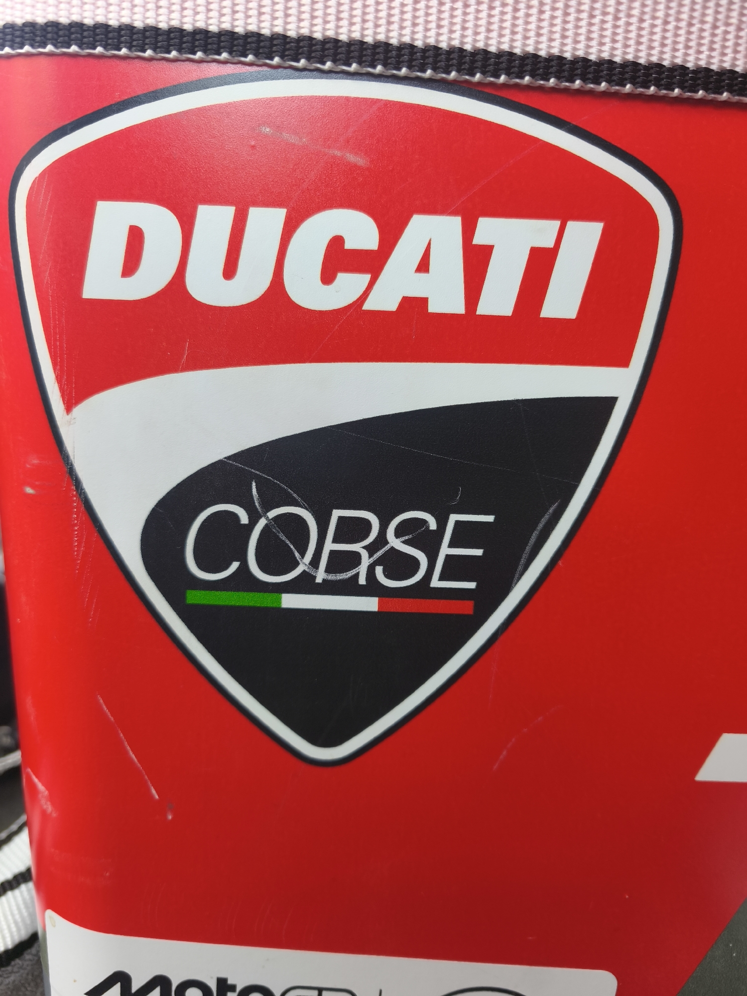 The image shows a close-up of a red object, likely a bag or piece of equipment, featuring the DUCATI CORSE logo. The logo is set against a red background with white and black accents. The DUCATI text is prominent in white lettering. Below it, the CORSE text is also in white. Underneath the CORSE text, there's a stripe of green, white, and red, representing the Italian flag. The object has visible scratches and scuffs, indicating wear and tear. The top edge of the object reveals a striped pattern in black and white. There is no visible indication of the city of Nàquera / Náquera, Spain within the image itself, nor any people or specific actions taking place.