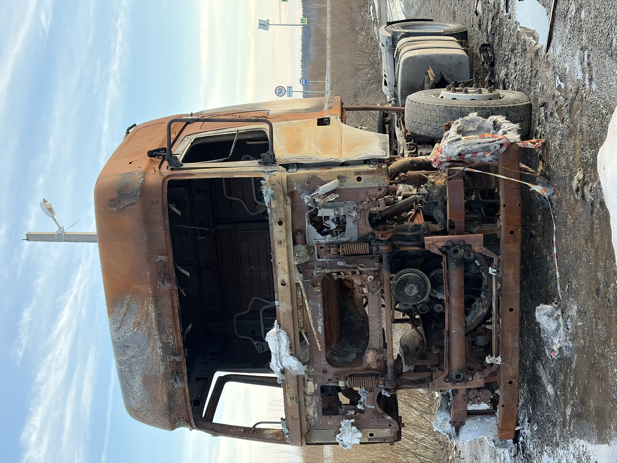 The image shows a severely burned truck cab beside a roadway. The location appears to be in a rural or semi-rural area. The truck cab is heavily damaged by fire, with most of the paint burned away and the internal components exposed. Parts of the engine and frame are visible, corroded and twisted by the heat. The wheels of the trailer are intact. The area around the truck is littered with debris and ash. Red and white barrier tape is present, indicating a potential hazard or crime scene. In the background, there is a roadway with trees and vegetation on either side. Roadside signs are visible in the far background, including one with speed limit 50 and an indication for a bus stop. The sky is partly cloudy. The presence of snow suggests a cold climate. The location is Zachernaya, Russia.