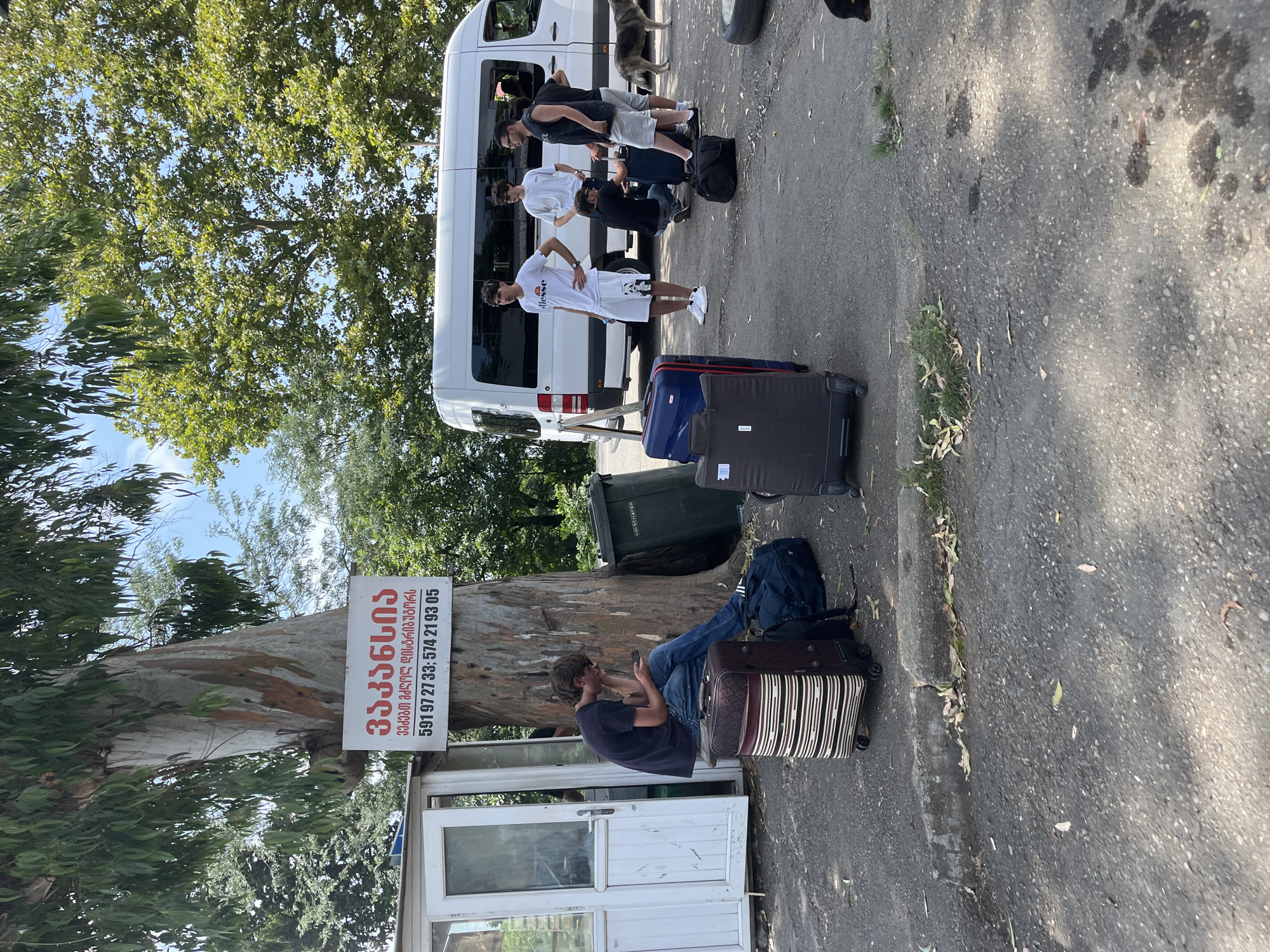 In Ureki, Georgia, a group of young men with luggage are gathered near a white van. One man, wearing a dark t-shirt and jeans, sits on a small stool next to a suitcase, looking at his phone. Several other men stand around the van, some with their hands on their hips, others attending to luggage. Suitcases, duffel bags, and a trash bin are visible on the asphalt ground. The scene is set against a backdrop of lush green trees and a bright blue sky, with a small white building to the left. A sign with Georgian text is affixed to a tree trunk.