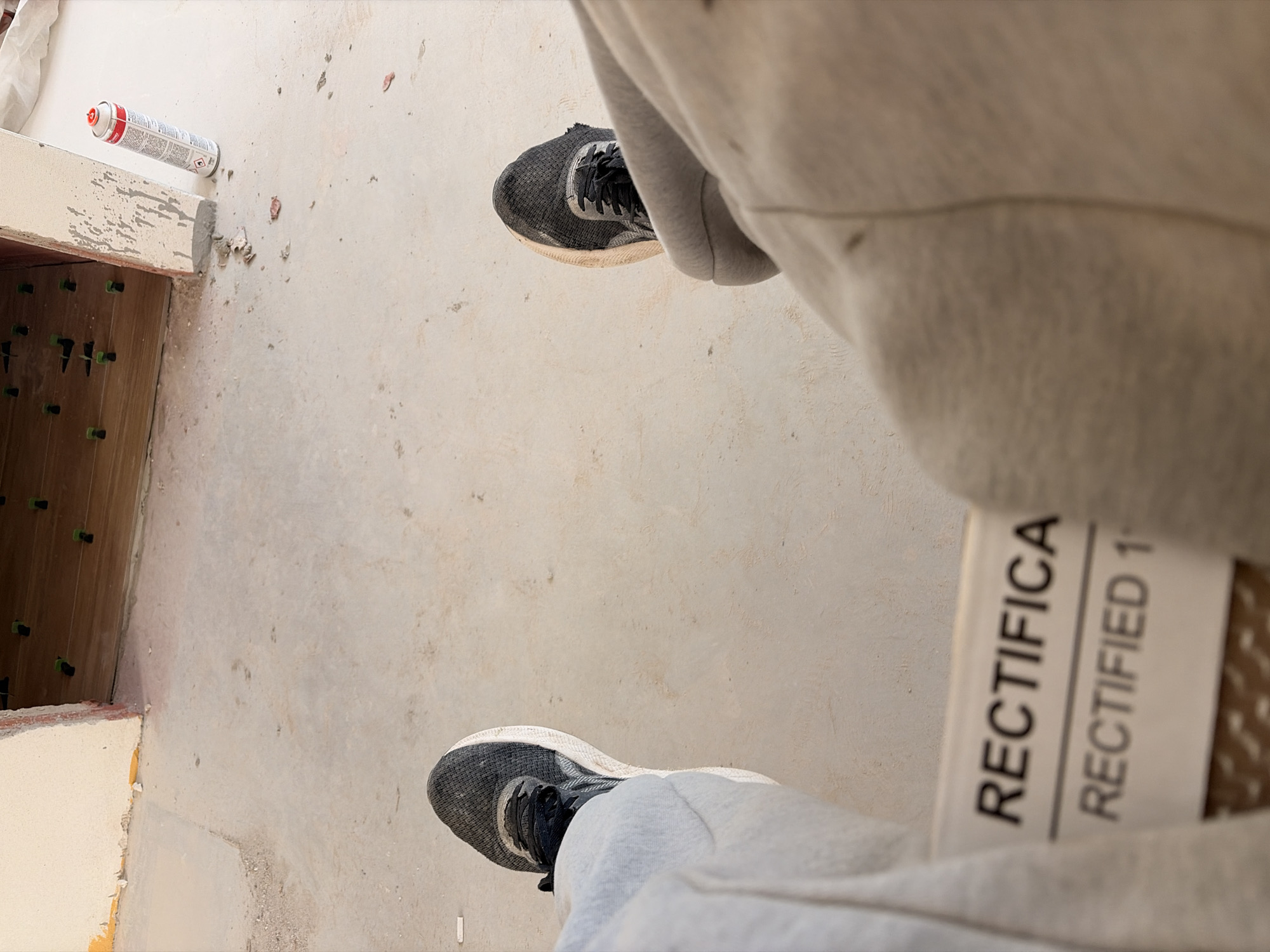 The image depicts a first-person perspective of a person's legs and feet, wearing athletic shoes and light-colored sweatpants, sitting on a surface. The environment appears to be an indoor construction or renovation site, characterized by a concrete floor with scattered debris. In the background, a partially finished wall with exposed construction materials and wooden flooring with green leveling spacers can be observed. A can of spray foam is also visible near the wall. A label on the person's lower leg displays the text "RECTIFICA" and "RECTIFIED 1". The specific location of Příbram, Czechia, is not discernible from the visual information provided in the image.