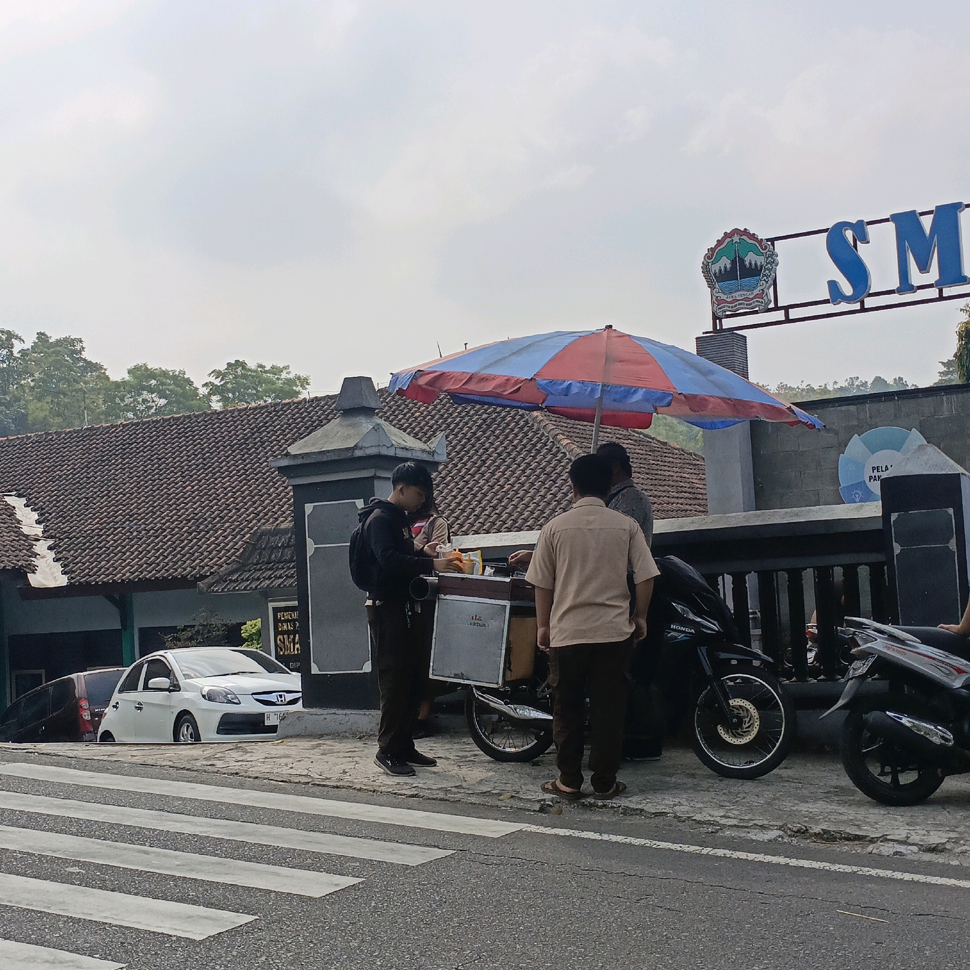A group of people are gathered near a food cart under a striped umbrella in front of a building in Sumogawe, Indonesia. The building has a tiled roof and a sign that reads "SMK" in large blue letters, suggesting it is a vocational high school. Two individuals in uniform shirts appear to be students, interacting with a vendor at the food cart. A black motorcycle is parked nearby, and a white car is visible on the left side of the image, parked behind a crosswalk. The sky is overcast, and trees can be seen in the background.