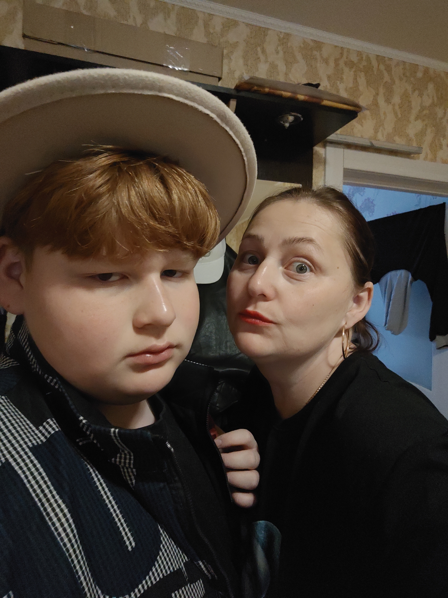 The photograph captures two individuals indoors. The person on the left is younger, wearing a light-colored wide-brimmed hat and a dark, patterned jacket. The person on the right, likely an adult, is wearing a black top, visible gold hoop earrings, and has red lipstick. The wallpaper behind them is patterned. A dark object is mounted on the wall above them, and a partially open doorway reveals clothing hanging inside. The photograph was taken in the city of Markovskii, Russia.
