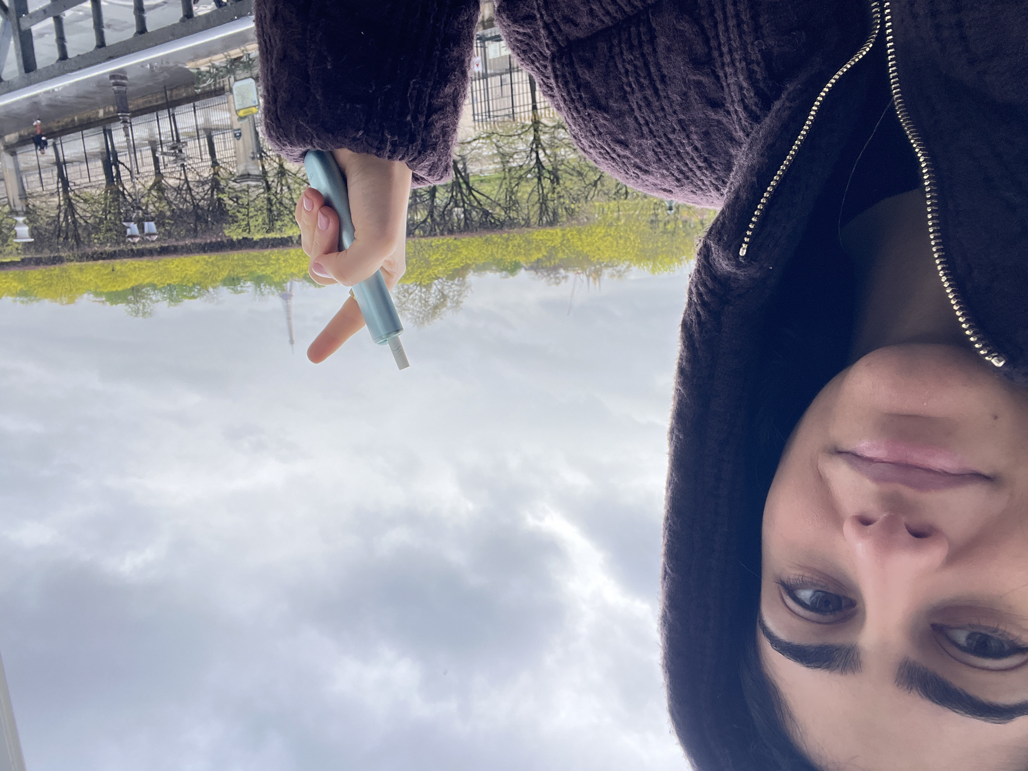 A female individual is depicted in a close-up, selfie-style photograph taken from an elevated position in Paris, France. The woman, with dark hair and eyebrows, looks directly at the camera with a slight smile. She wears a dark-colored, textured, hooded jacket with a visible silver zipper. Her right hand, featuring light pink nail polish, holds a light blue-green electronic device, possibly a heated tobacco product, from which a white cylindrical component protrudes.
The background reveals a city street appearing wet, suggesting recent precipitation, viewed over a dark wrought iron railing. Beyond the street lies a park or garden characterized by trees with vibrant green spring foliage. The Eiffel Tower is distinctly visible in the far distance, silhouetted against an overcast sky. Additional elements include street lampposts, a metal fence, and a distant pedestrian wearing a red top. The ambient light is diffused, consistent with a cloudy daytime.