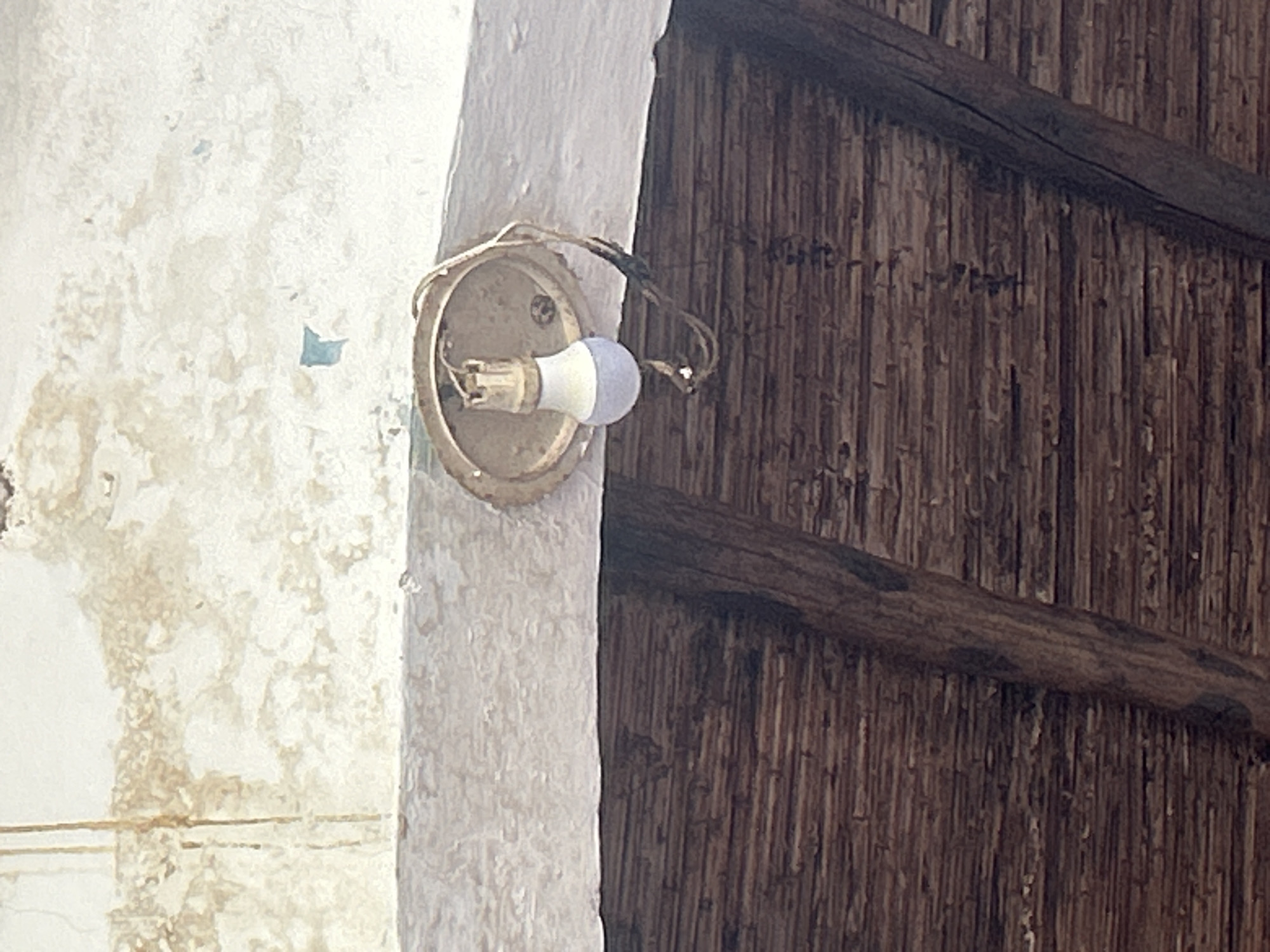 A close-up, low-angle shot captures a damaged outdoor light fixture affixed to a white, weathered overhang. The light bulb, partially unscrewed, hangs precariously from its socket, wires exposed. Below the overhang, a rustic ceiling constructed of parallel wooden beams and woven reeds suggests an open-air or covered patio area. The upper portion of the image reveals a stained and peeling white ceiling, possibly indicative of water damage, with smudges of brown and faint blue. The scene appears to be in an area with a tropical or sub-tropical climate, possibly in Santa Ana, Venezuela, given the architectural style and weathered materials. There are no people or animals present.