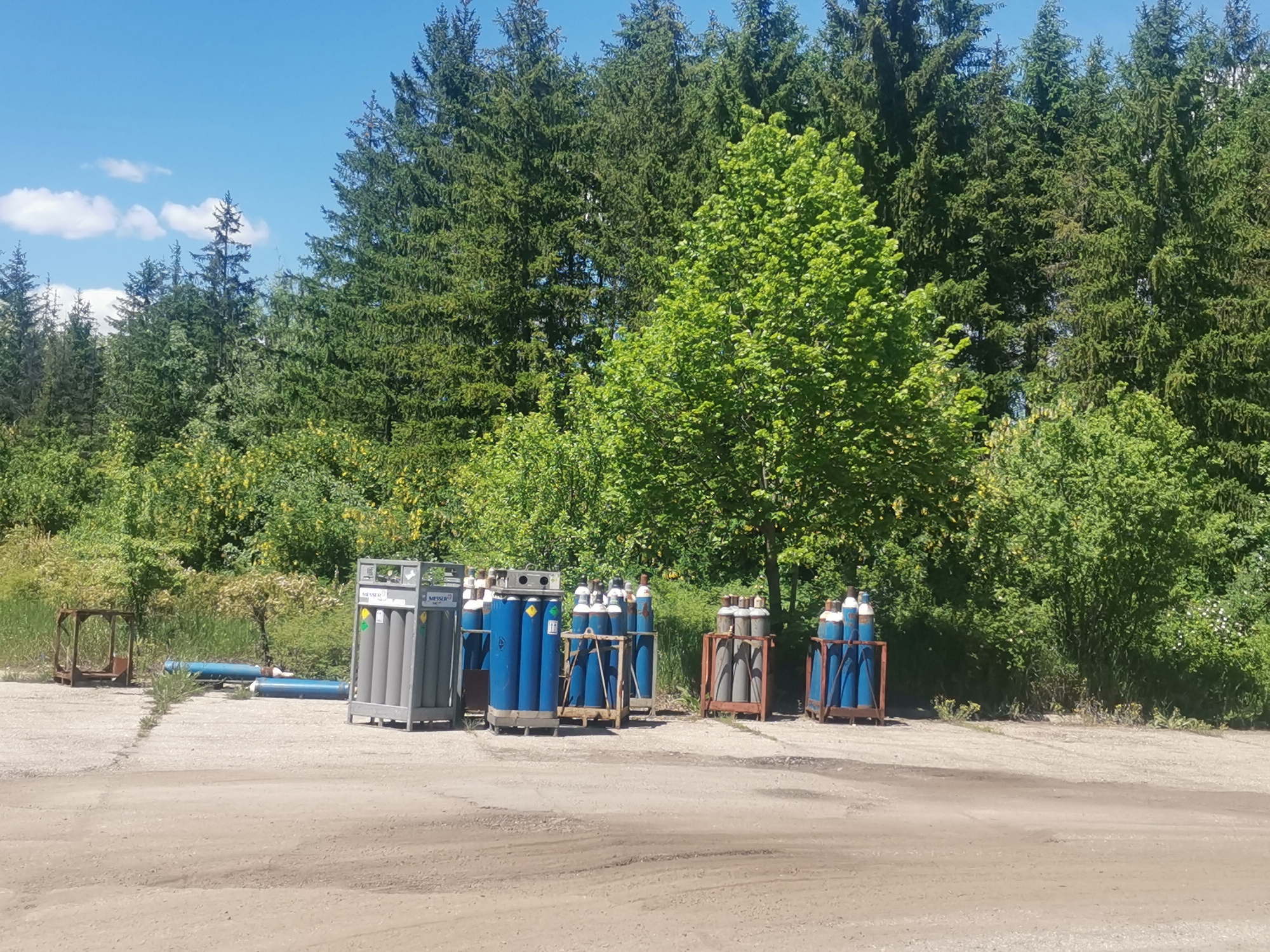 The image depicts a storage area in Zhivkovo, Bulgaria, likely an industrial or commercial zone. Several metal racks filled with blue and gray gas cylinders are arranged on a dirt and gravel surface. In the background, a dense forest of pine and deciduous trees rises behind the storage area. A clear blue sky with scattered clouds is visible above the trees. Additional metal frames and blue pipes are present on the left side of the image. The scene is static and appears to be outdoors during daylight hours.