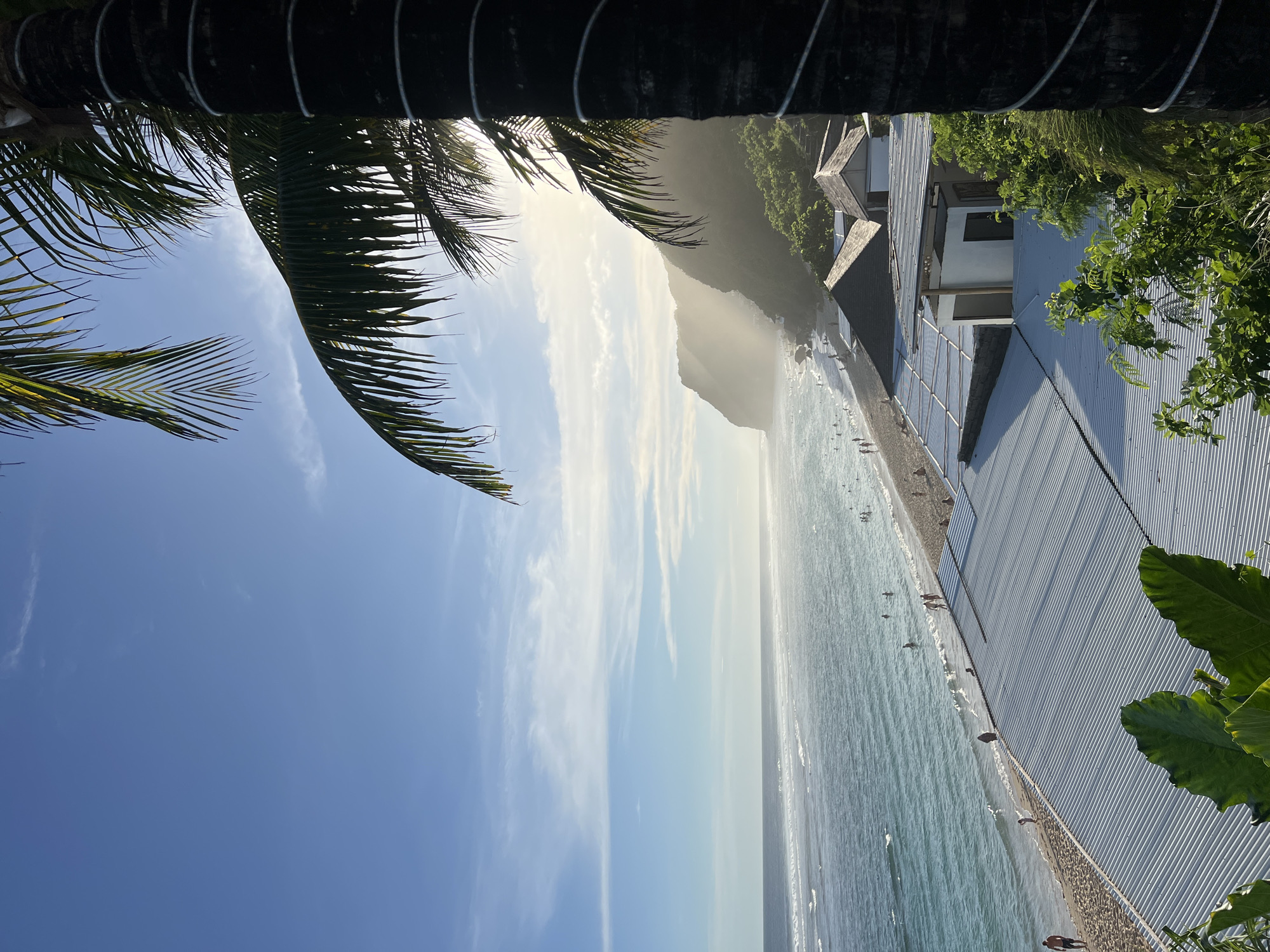 This image presents a picturesque tropical beach scene, likely captured during late morning or early afternoon due to the bright, high sun. A prominent palm tree trunk and dark fronds frame the upper left portion, silhouetted against a clear, light blue sky with scattered wispy clouds.
In the mid-ground, a wide expanse of ocean stretches towards the horizon, exhibiting calm, turquoise waters with gentle waves breaking near the shore. Numerous individuals are visible in the water and on the sandy beach, engaging in leisure activities such as swimming, wading, or simply relaxing by the sea.
To the right, a series of modern beachfront buildings with distinctive corrugated metal roofs and facades line the shore. Lush green vegetation, including large leafy plants and denser foliage, thrives around these structures and extends up the slopes of a large, mist-shrouded mountain or hill that dominates the background. The sun's intense light creates a bright haze around the distant mountain and sky, adding to the serene and inviting atmosphere. The scene suggests a popular coastal destination, consistent with Kuta Selatan, Indonesia, known for its beaches and natural beauty. No discernible text is visible within the image.