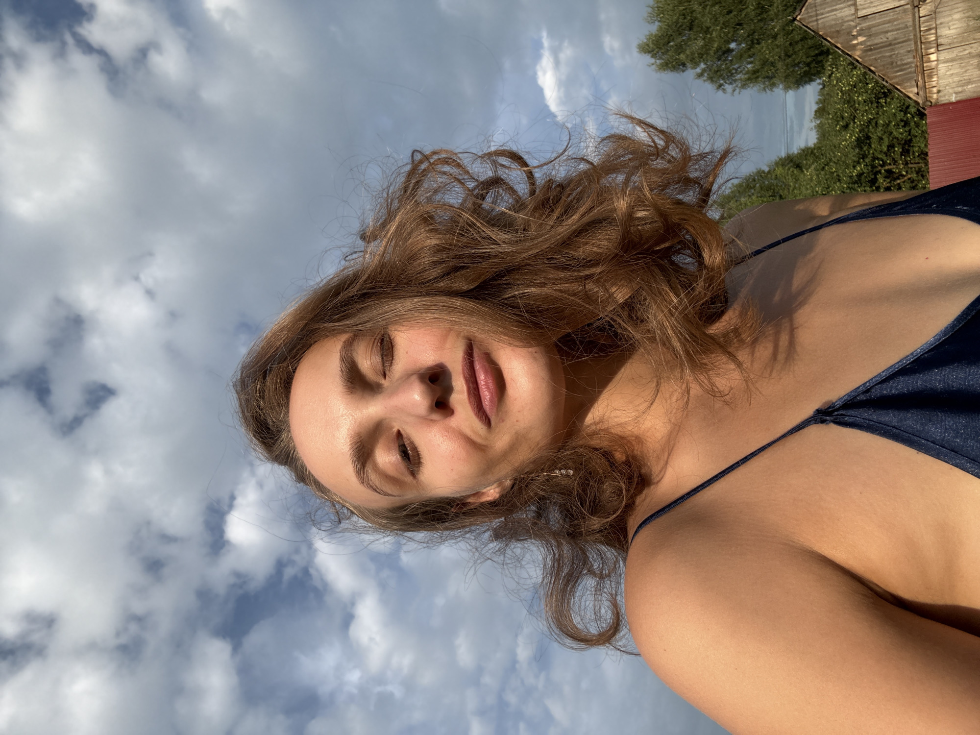 A young woman with auburn, wavy hair is pictured in a close-up shot outdoors, with a cloudy sky as the backdrop. She is wearing a dark blue, spaghetti-strap garment. Her head is tilted slightly, and her eyes are partially closed, with a subtle smile on her lips. The lighting suggests it is either early morning or late afternoon, casting a warm glow on her skin. The background features green trees and a rustic wooden structure, consistent with a rural setting like Sharkan, Russia.