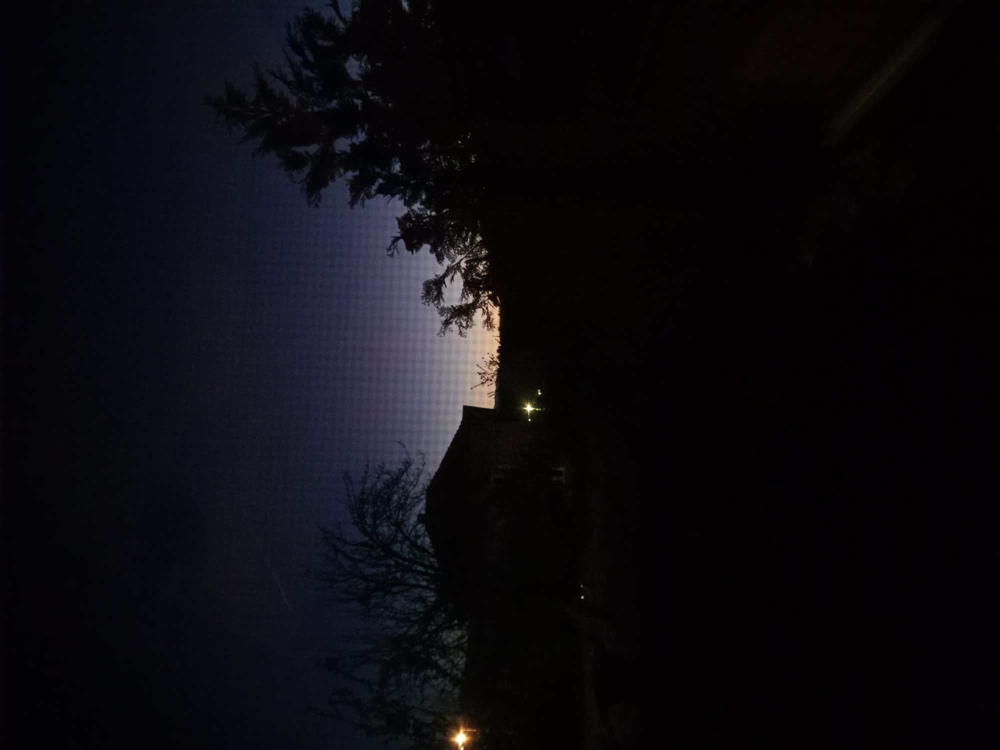 The image depicts a night scene in an unknown city. A dark building, possibly a house or church, is silhouetted against a partially illuminated sky, with a tree in the foreground. Lights are visible within the building and outside. The image is dark and obscured, making it difficult to discern further details.